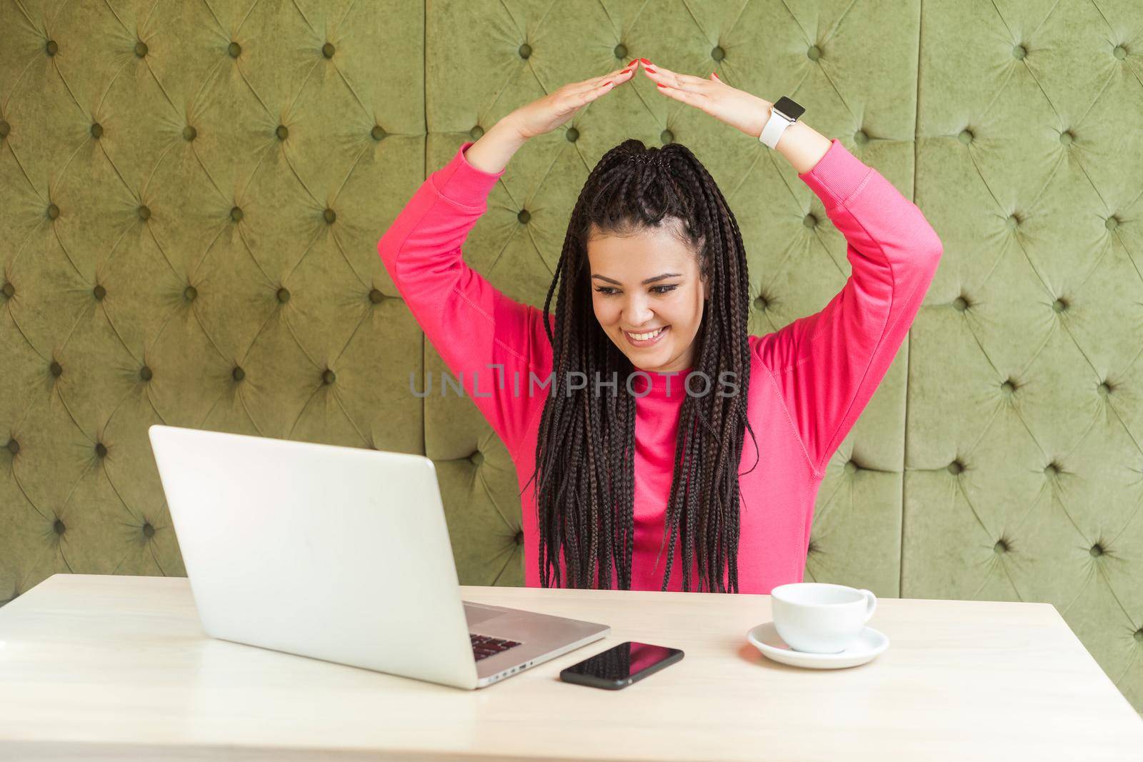 I'm in safety. Portrait of childish young girl with black dreadlocks hairstyle in pink blouse are sitting in office and holding hand upper hand, making roof with hands, looking at laptop, toothy smile