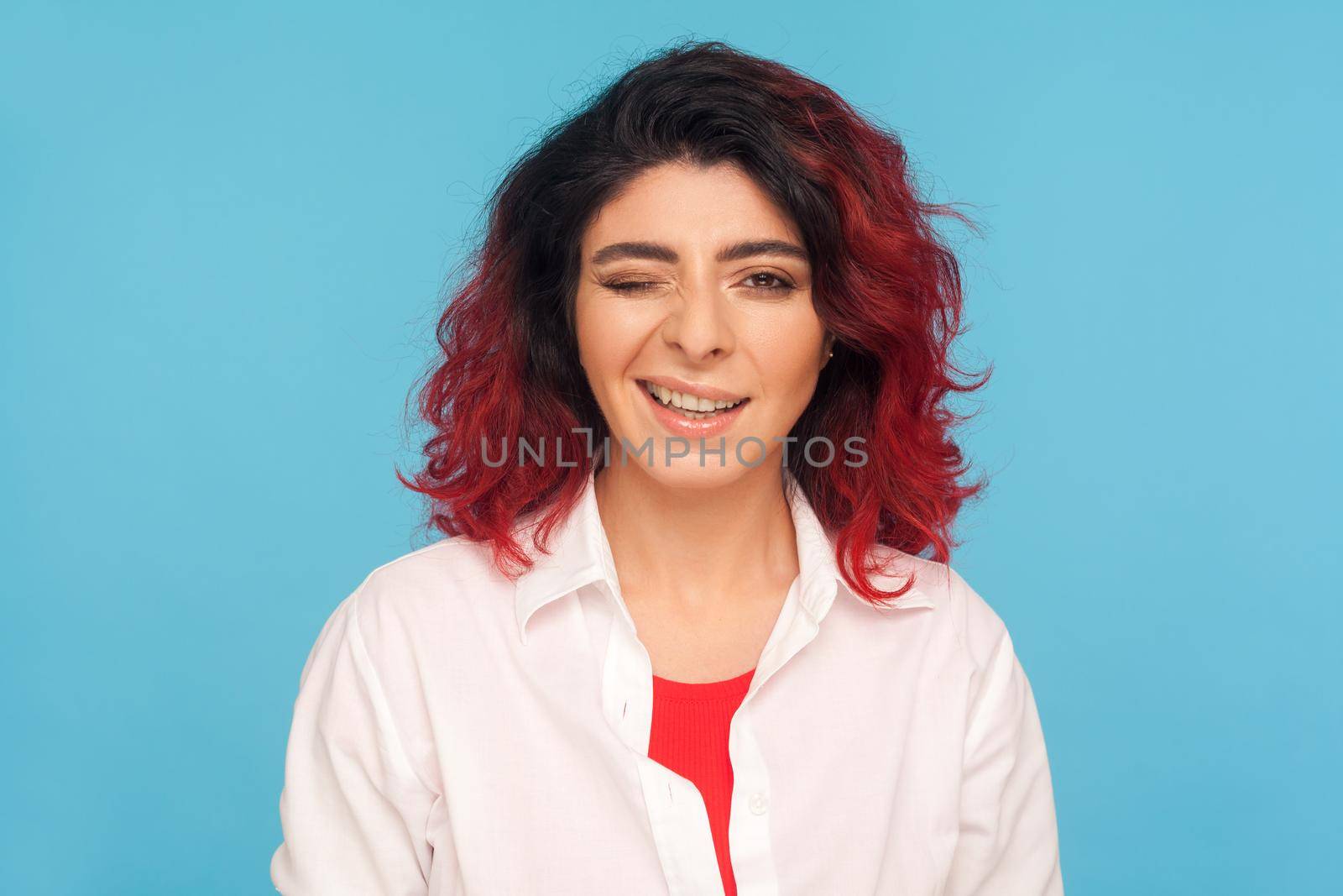 Portrait of charming playful hipster woman with fancy red hair in white shirt winking to camera and smiling, cheering up with happy look, having interesting idea. indoor studio shot, blue background
