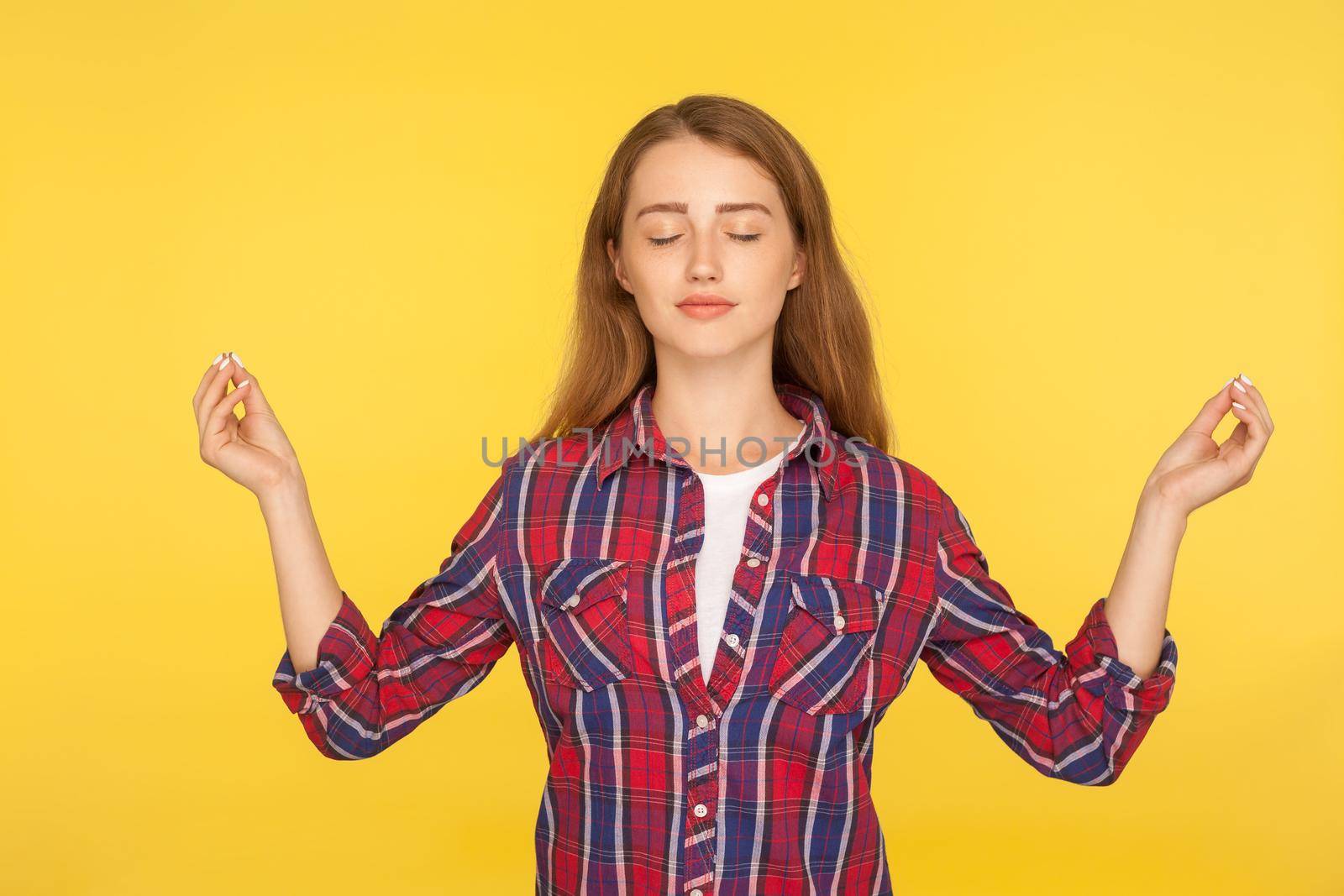 Harmony and inner peace. Portrait of happy calm ginger girl in checkered shirt practicing yoga, holding hands in namaste gesture, meditating with closed eyes. studio shot isolated on yellow background