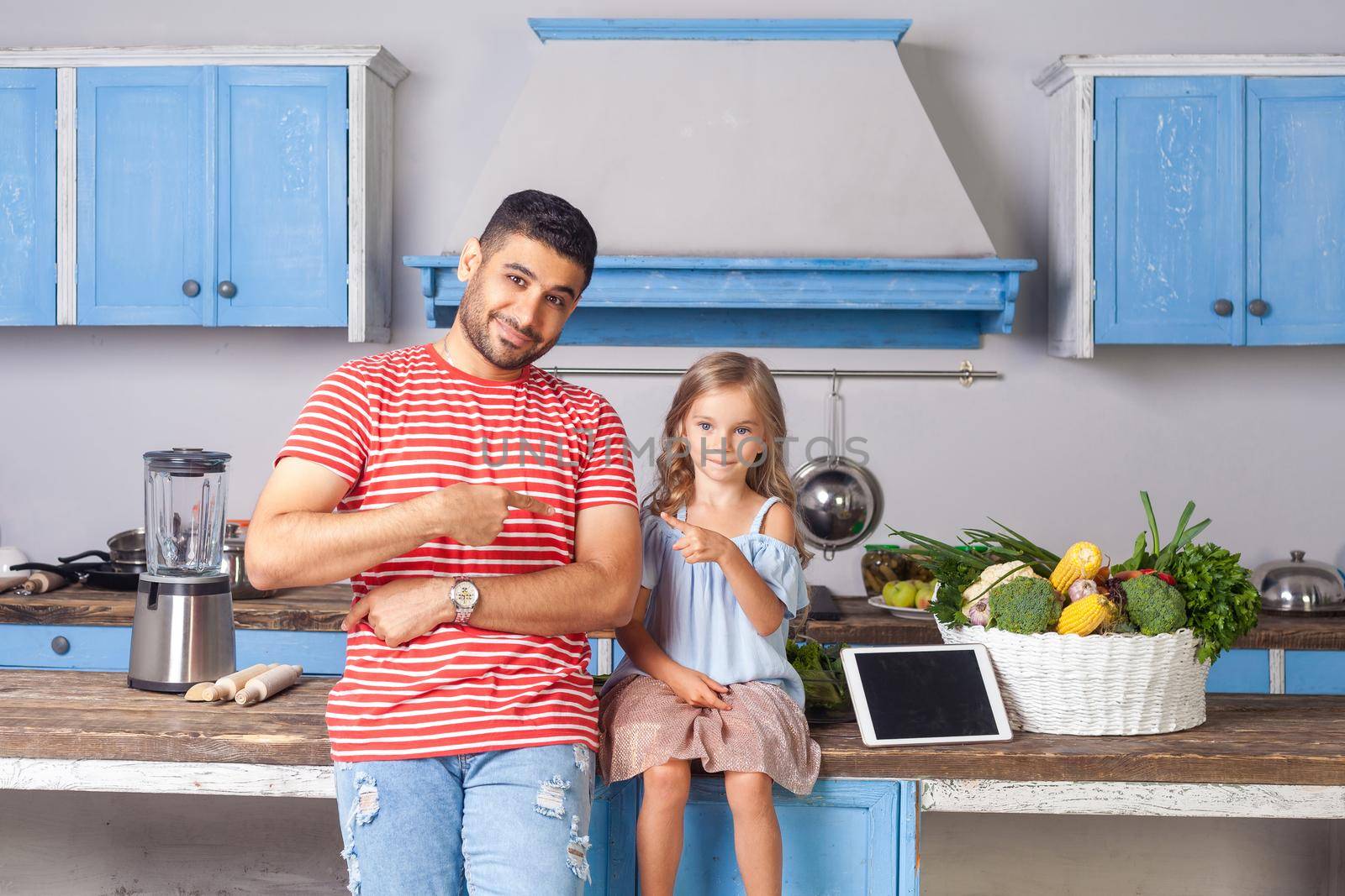 Your turn to cook. Father and daughter pointing each other and smiling at camera, sitting in modern kitchen, tablet pc and vegetables on table behind, recipes from internet, vegetarian nutrition, diet
