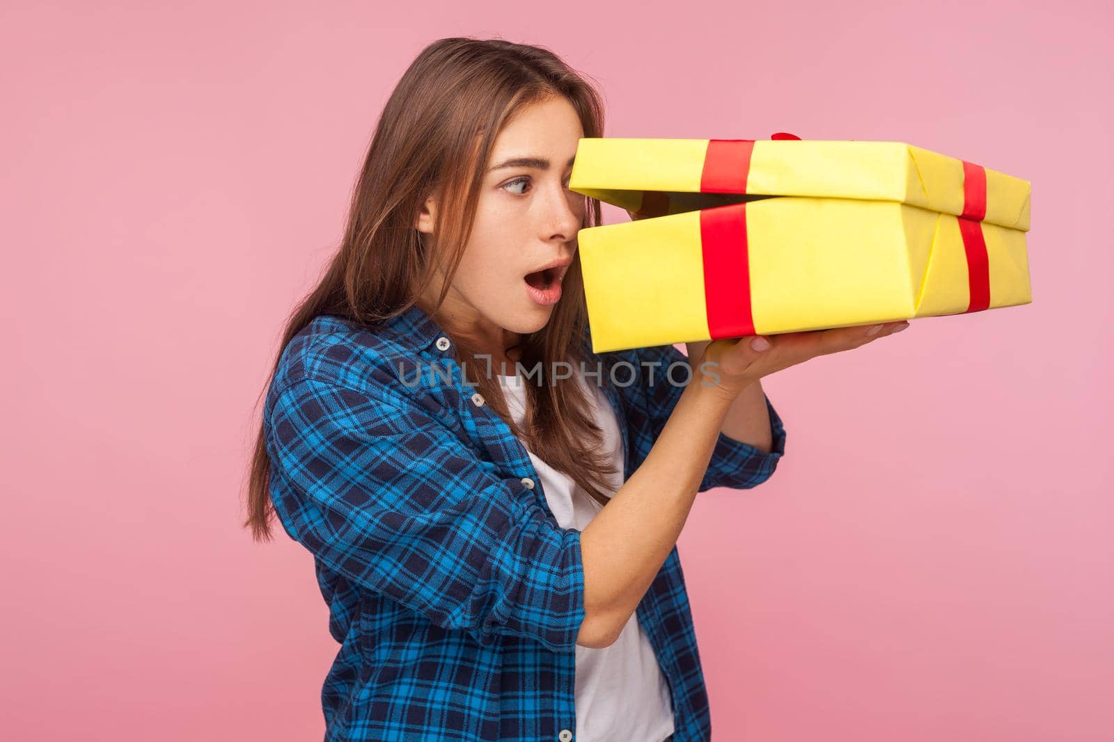 Portrait of curious amazed girl in checkered shirt looking inside present box with astonished shocked expression, pleasantly surprised by birthday gift. indoor studio shot isolated on pink background