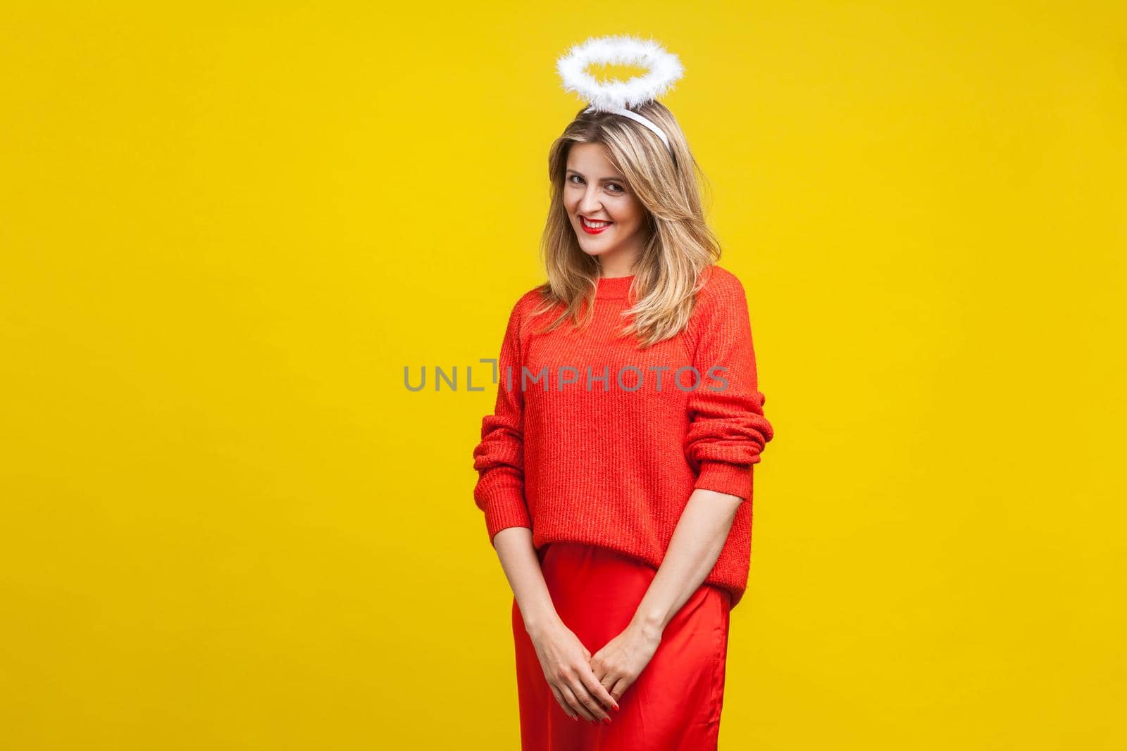 Portrait of modest angel beautiful young woman with bright makeup in red casual clothes standing with halo on her head and looking shy at camera. indoor studio shot isolated on yellow background