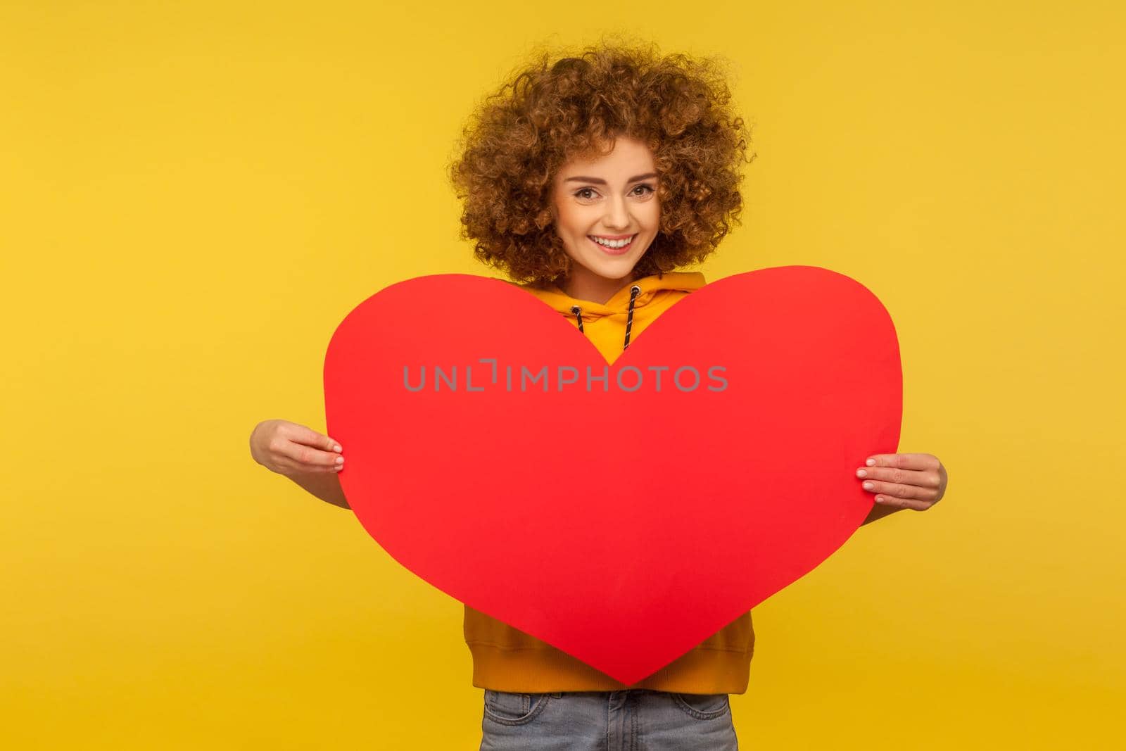 Charming lively energetic woman with fluffy curly hair holding big heart love symbol, looking at camera with toothy smile, romance on Valentine's day. indoor studio shot isolated on yellow background