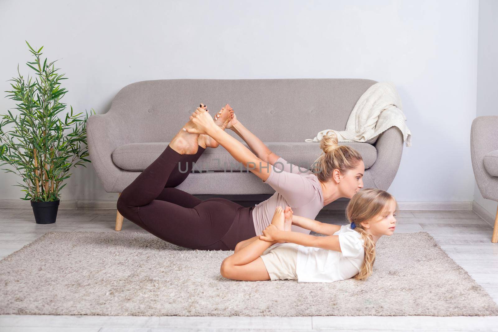 Sportive young mother and charming daughter practicing yoga at home, standing in bow pose, happy family doing gymnastics exercise together, fitness training with child. body care, healthy lifestyle