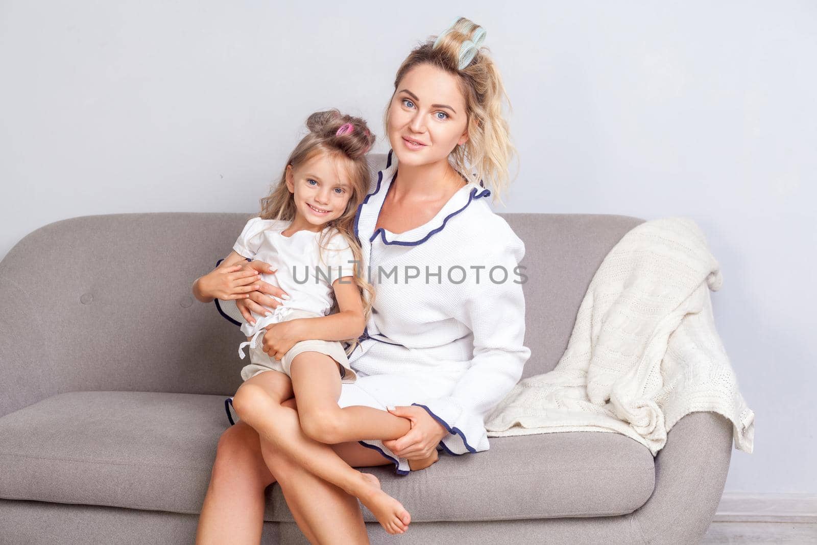 Beautiful happy mother and cute daughter with curlers in hair sitting on sofa and smiling looking at camera, spending time together at home, great parenting, motherhood, family communication. indoor