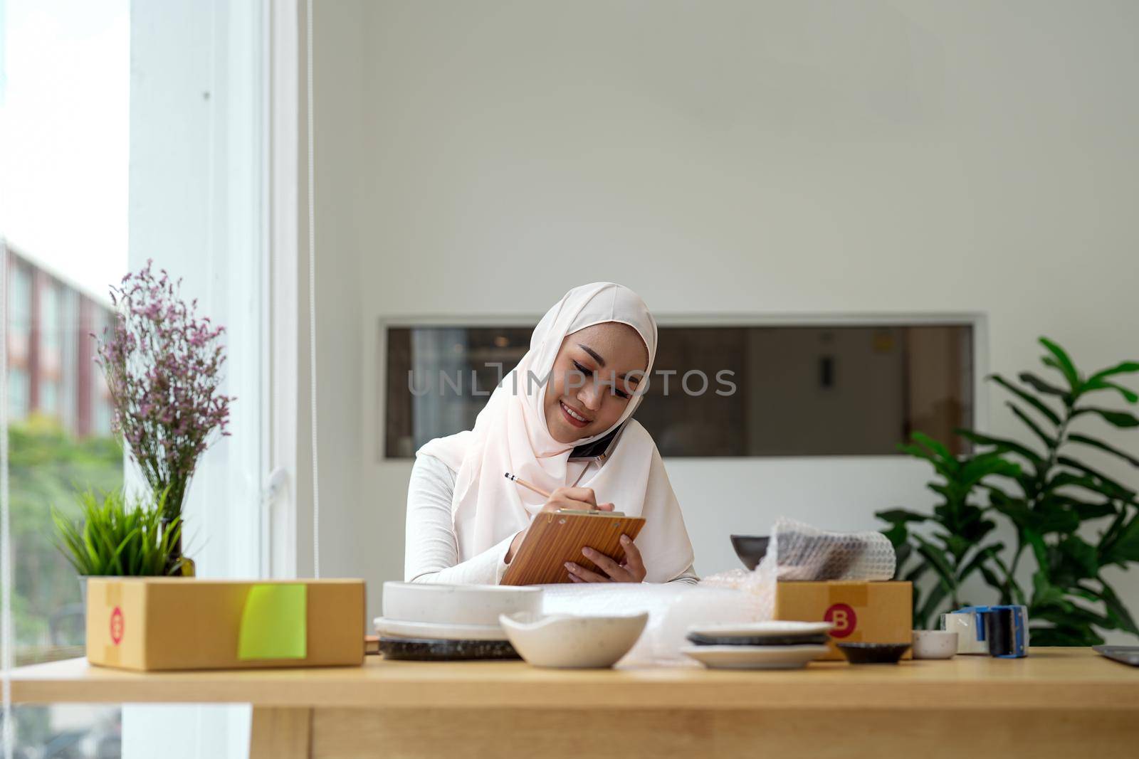 Young asian muslim woman business owner with many parcel boxes on the table happy online sales job, calling with smartphone, get an order from customers, take notes, and make arrangements for delivery by post