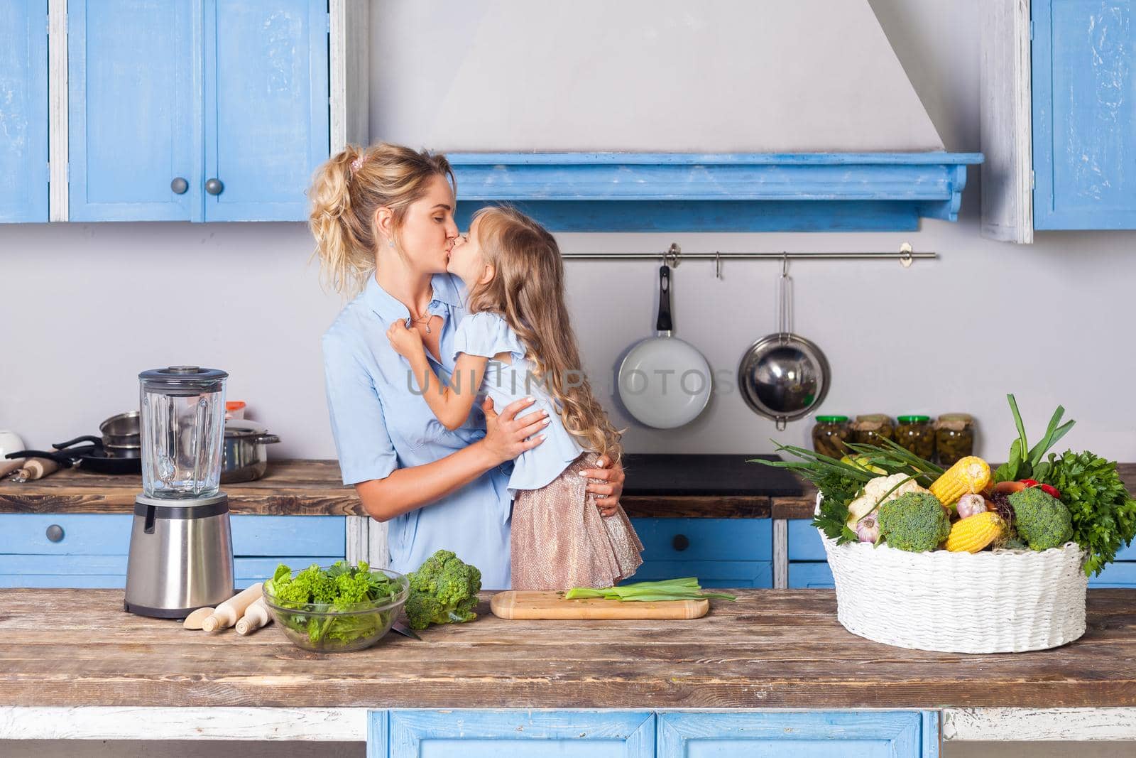 Lovely daughter kissing young mother while cooking together in kitchen, preparing salad for breakfast, basket of fresh green vegetables and blender on table, vegetarian food, healthy nutrition