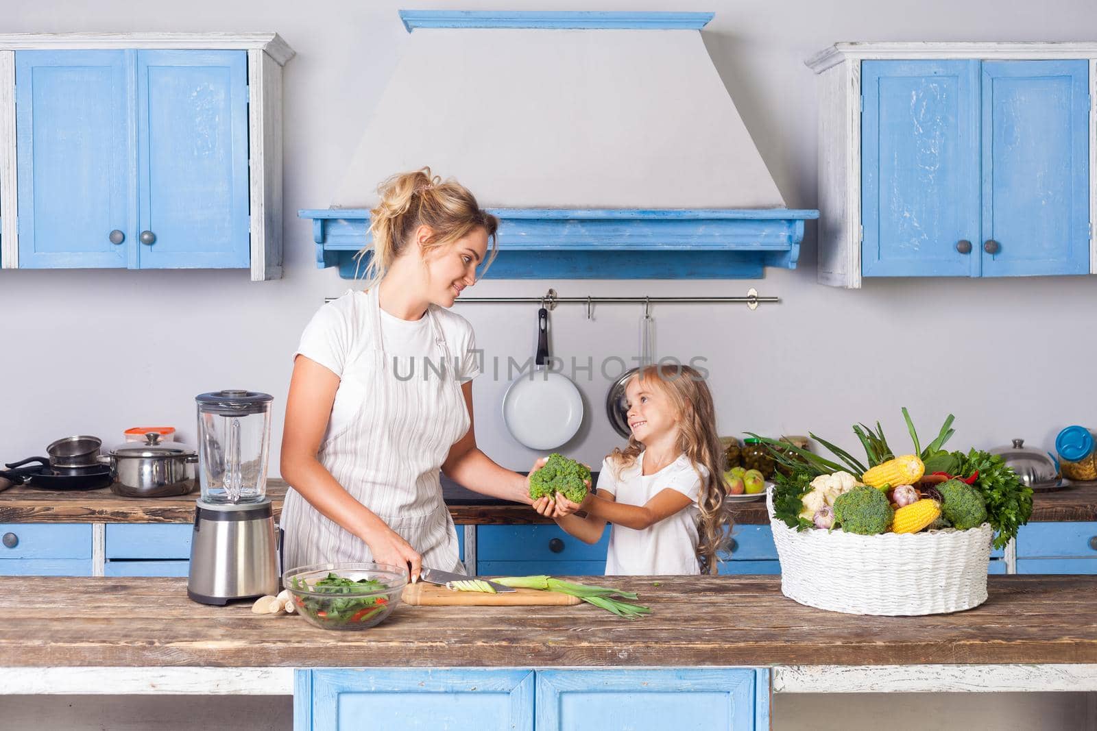Happy cute mother in apron and little daughter holding broccoli and smiling, cooking salad in kitchen together, preparing vegetarian food, basket of fresh vegetables on table, healthy vegan nutrition