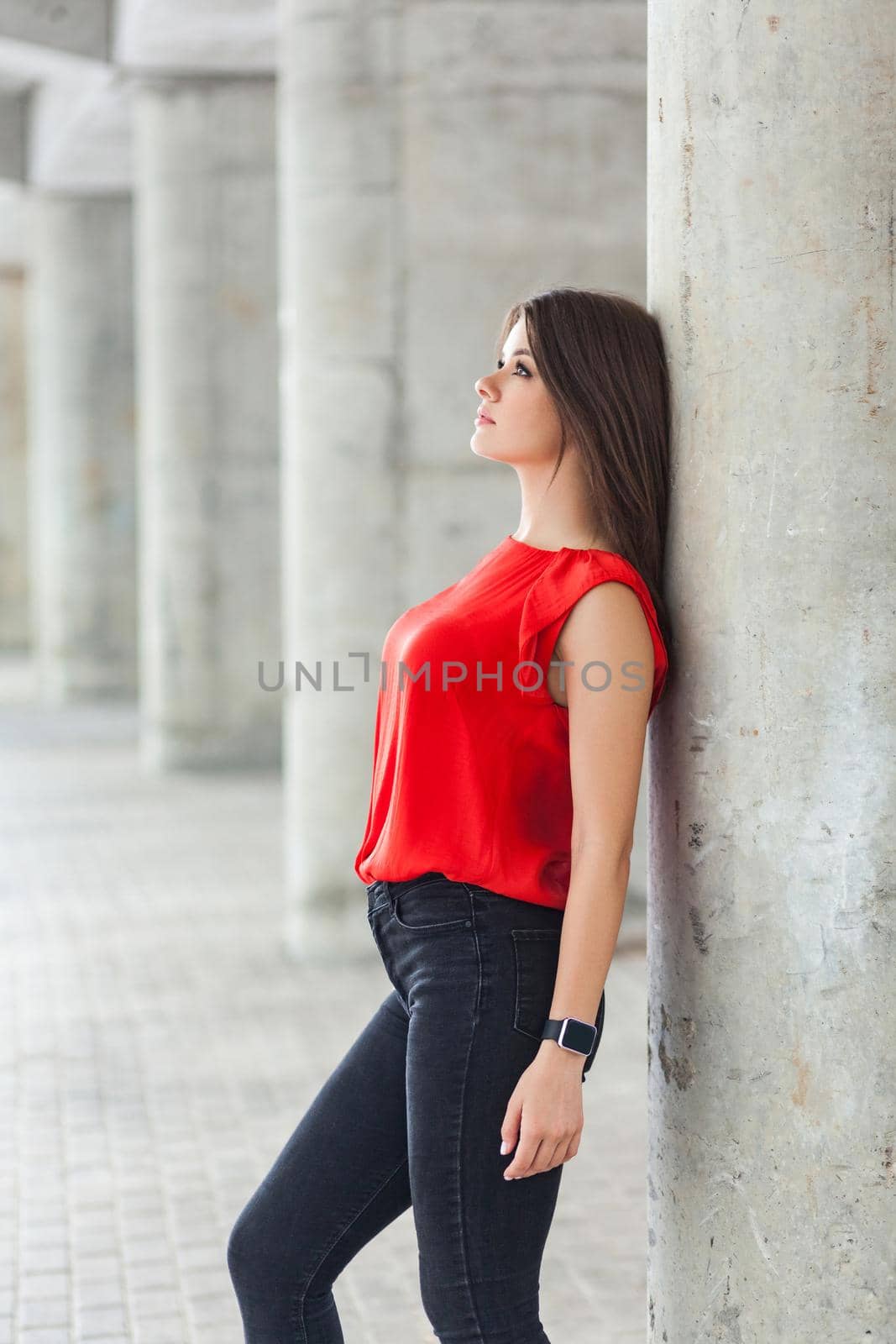 beautiful woman in red blouse and jeans posing while leaning on gray column. outdoor shot.
