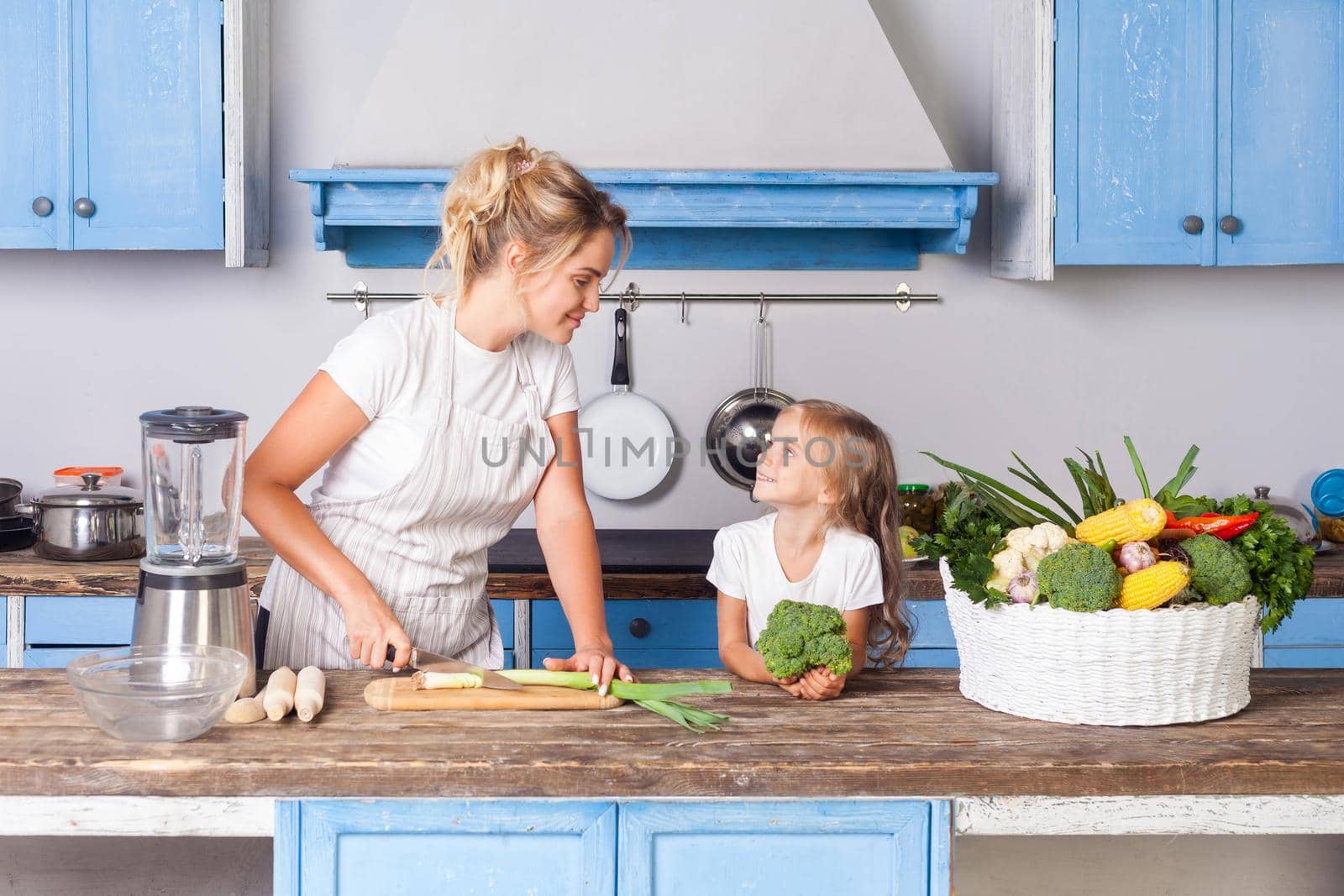 Happy pretty mother in apron chopping cutting green vegetable with knife and smiling to little daughter, woman cooking salad in kitchen, preparing vegetarian food, basket of fresh vegetables on table