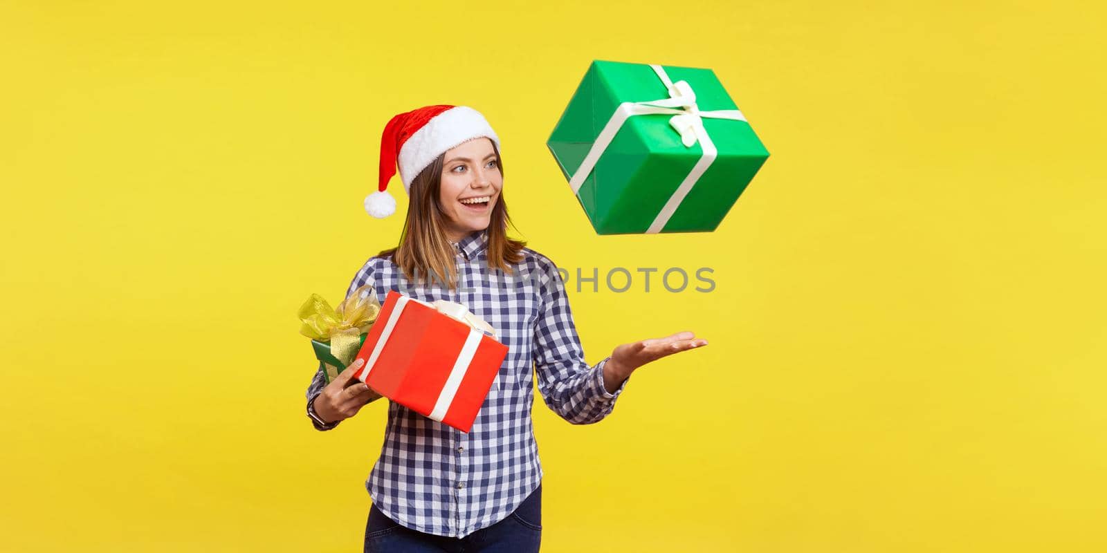 Portrait of happy brunette young woman in santa hat and checkered shirt standing throwing gift boxes in air or catching presents, christmas shopping. indoor studio shot isolated on yellow background