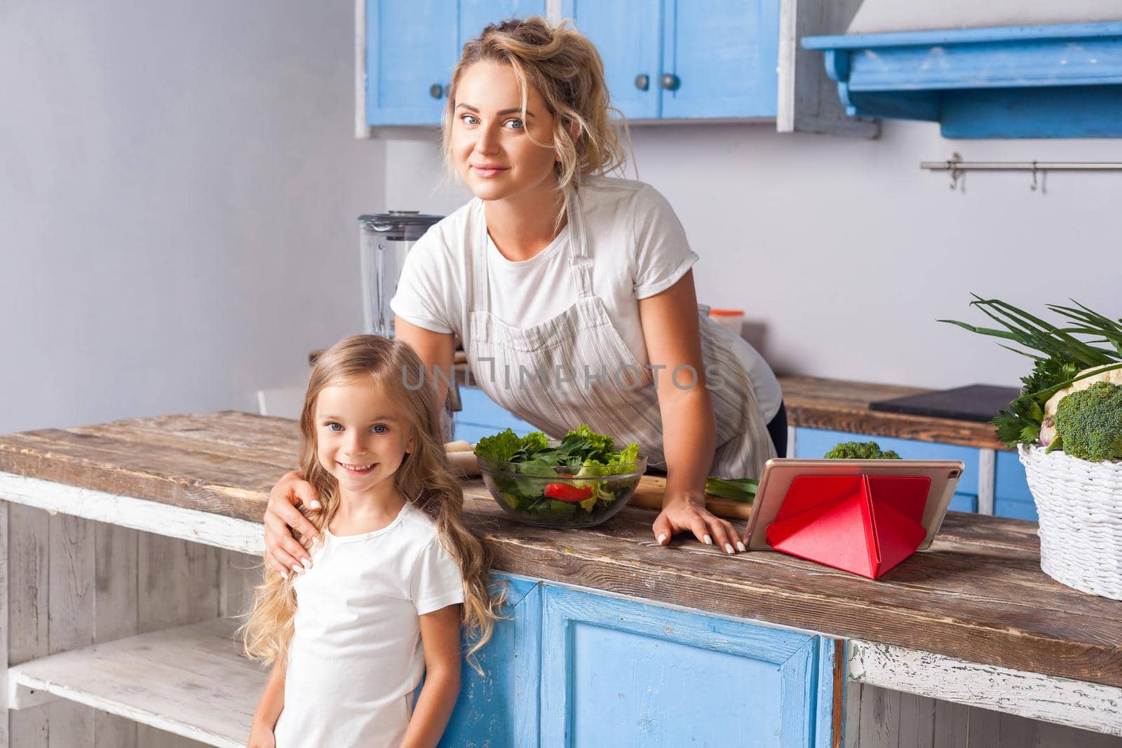 Cute woman and her daughter smiling at camera standing in modern kitchen, mother keeping hand on girl's shoulder, cooked salad with fresh green vegetables on table, vegetarian food, healthy nutrition