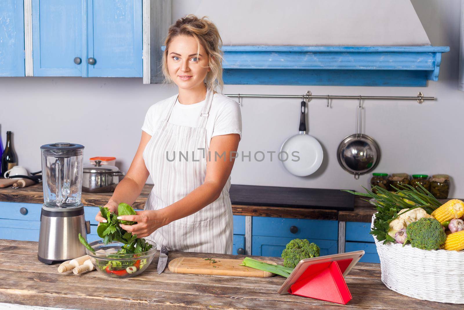 Attractive young woman in apron cooking green salad, preparing vegetarian food in modern kitchen and looking at camera, blender and basket of fresh vegetables on table, vegan diet, healthy nutrition