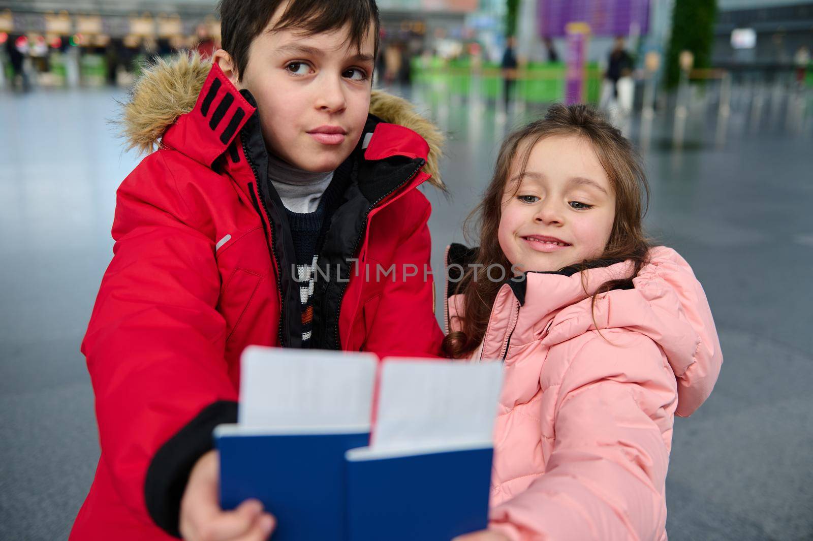 Adorable Caucasian kids- boy and girl, siblings holding boarding pass and Identity document in outstretched hands while waiting for check-in flight in the departure hall of an international airport