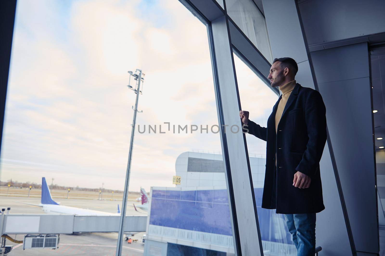 Full length portrait of a confident man passenger looking through panoramic windows overlooking the runway in the departure terminal of an international airport while waiting for a flight boarding