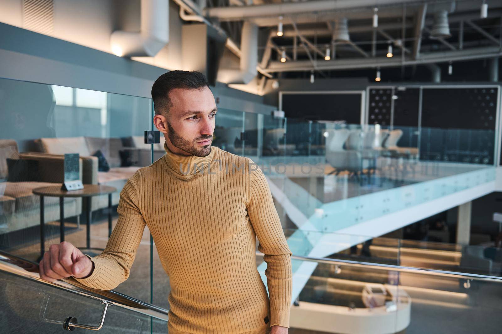 Handsome confident young Caucasian man in casual attire, confidently looking away while walking up stairs in VIP lounge in international airport departure terminal, awaiting the flight boarding