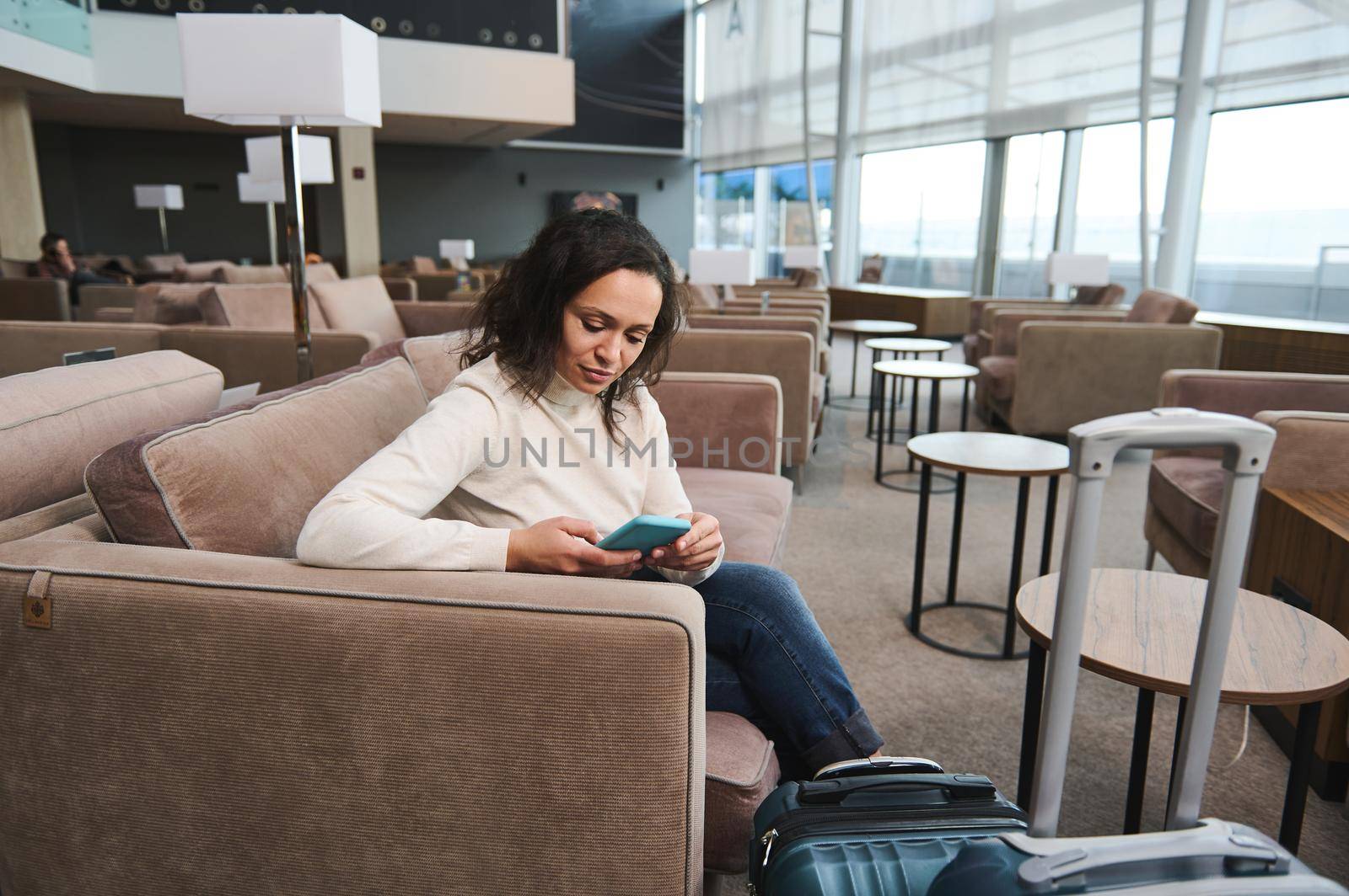 Beautiful young Middle-Eastern woman in casual wear using mobile phone, browsing websites, booking hotel while resting on a comfortable sofa in the VIP lounge, awaiting flight at international airport