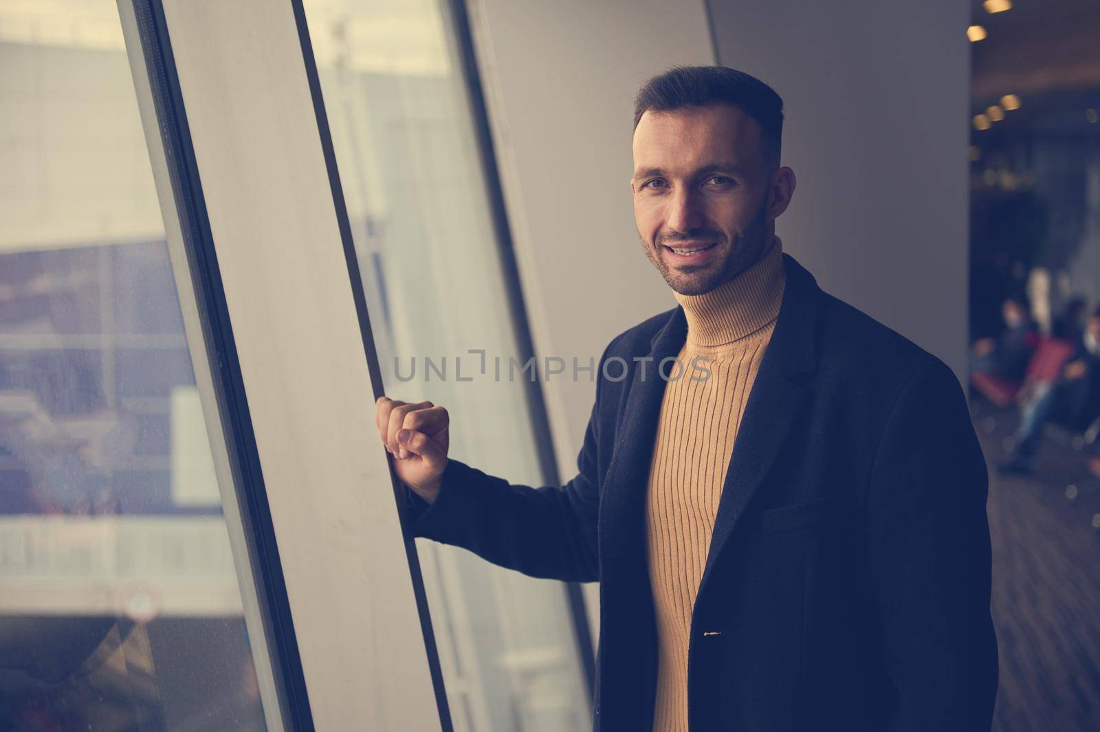 Toned photo, portrait of a handsome young man, transit passenger, traveler, smiling at camera, standing near panoramic windows overlooking runway in the departure terminal of international airport