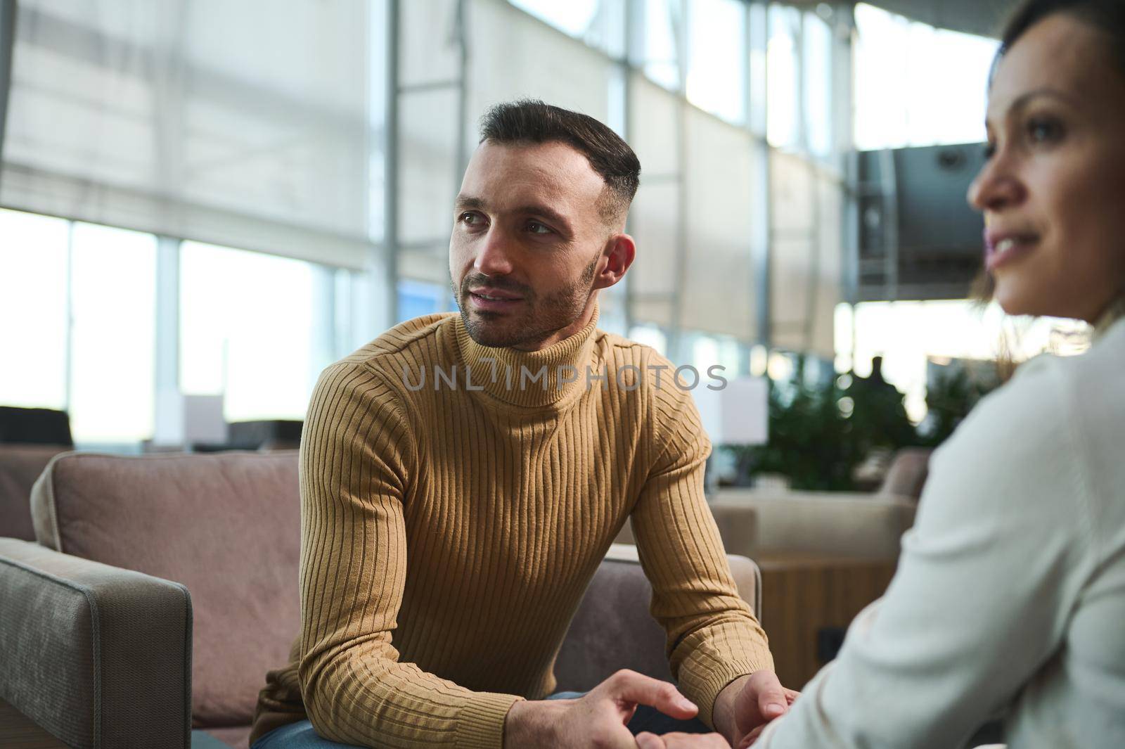 Focus on handsome Arabic man in pullover looking away, holding hands of his girlfriend, sitting together in chair in VIP lounge at international airport against panoramic windows overlooking runway