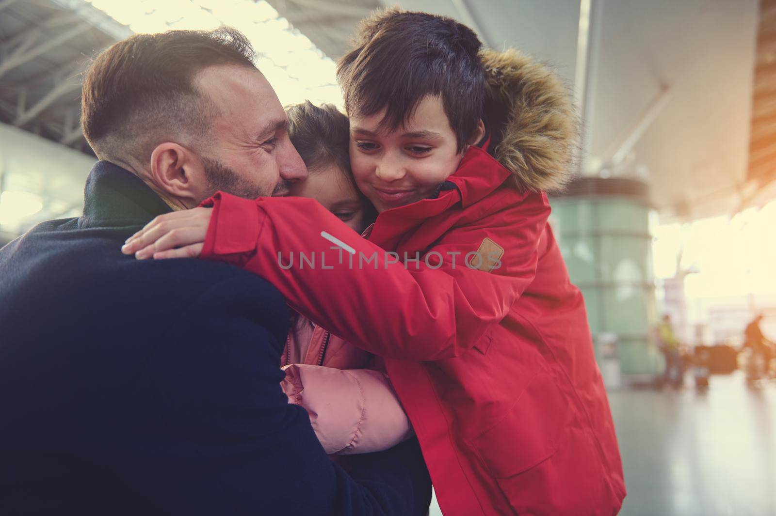 Emotional portrait of a happy young Caucasian man - father hugging his children - son and daughter in the international airport terminal. Long awaited family meeting and reuniting concept