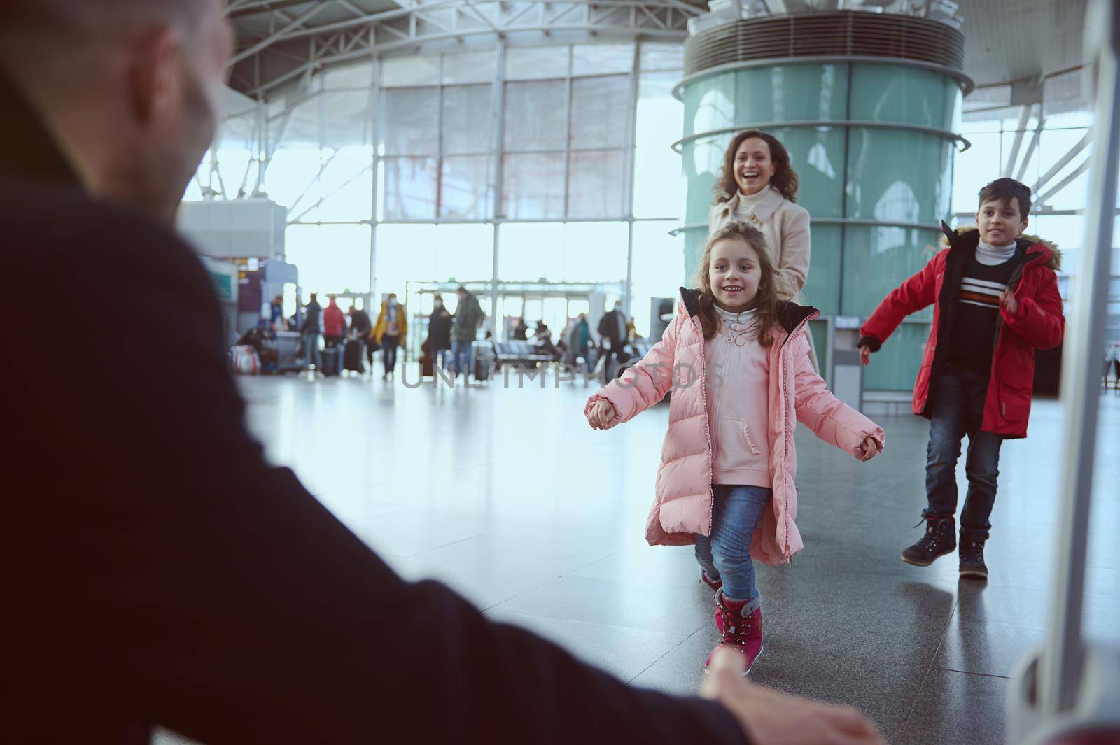 Happy excited woman with her kids running to her husband in the airport arrivals terminal waiting room, meeting him after his business trip. Long-awaited family reunion concept