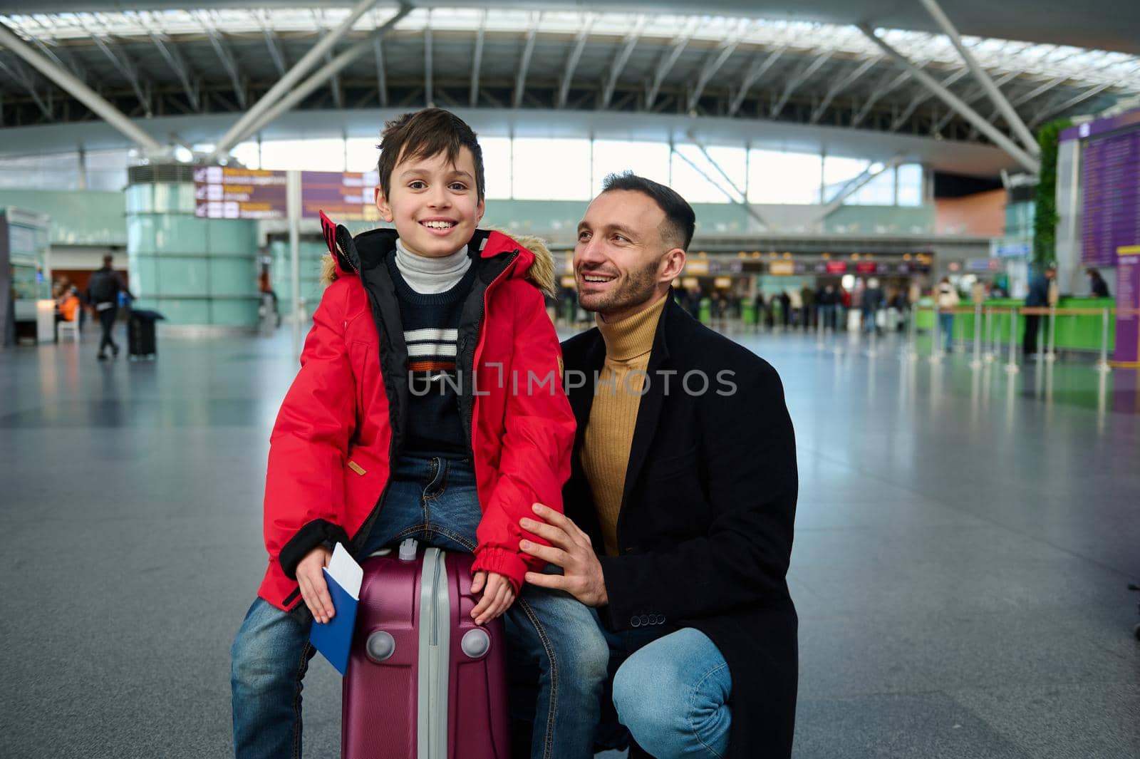 Handsome adorable preschooler, Caucasian child boy in warm red down jacket holding boarding pass and passport sitting on suitcase next to his loving father in international airport departure terminal