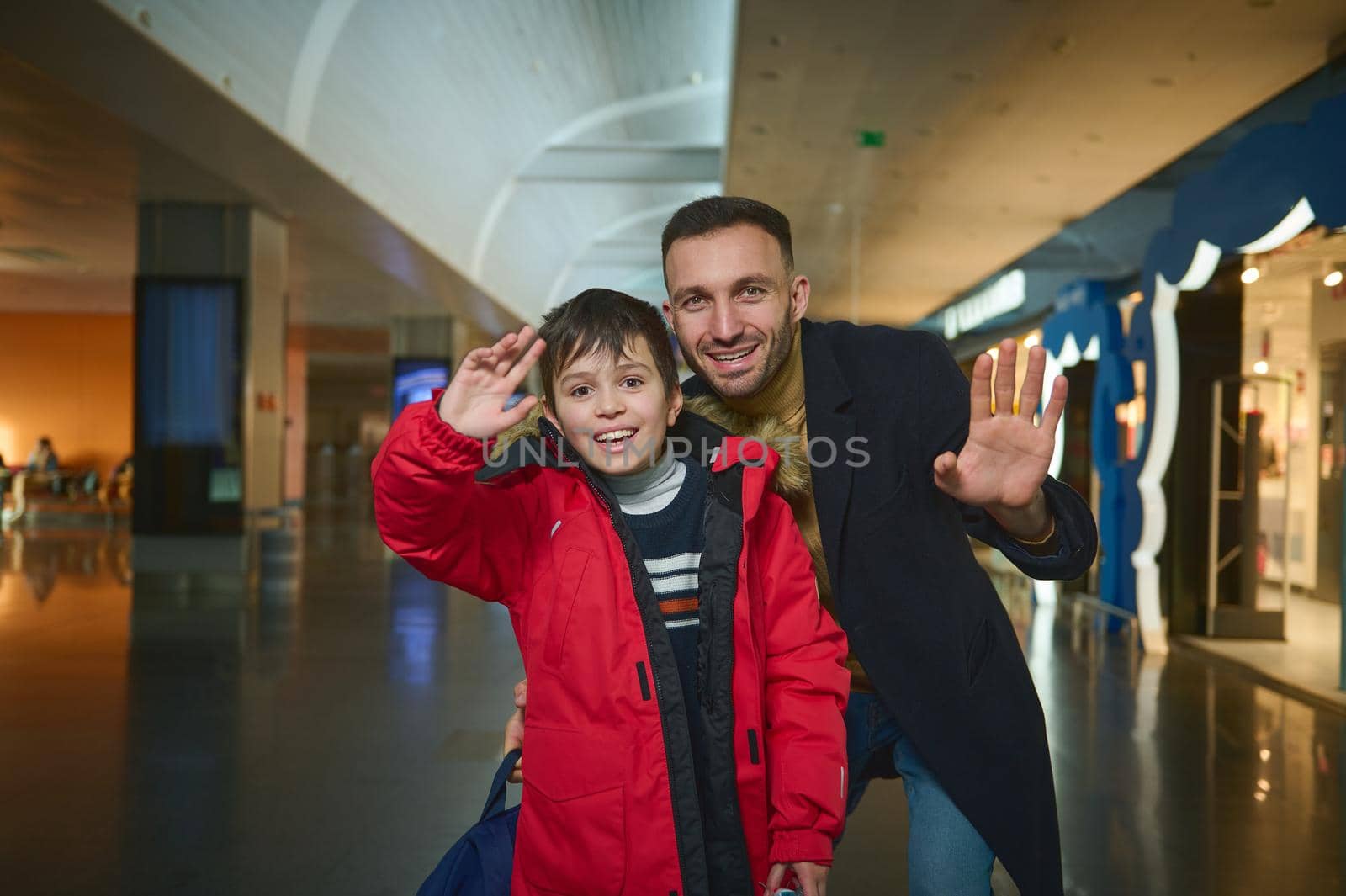 Happy Caucasian boy and his cheerful handsome dad, transit passengers smiling and waving looking at camera while walking along duty free stores in the departure terminal of an international airport