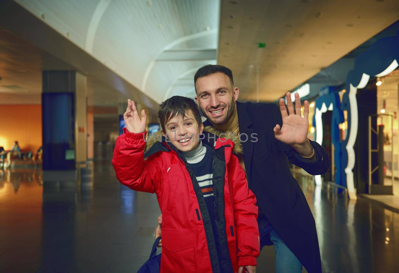 Handsome Middle Eastern man father and his adorable son, passengers, waving looking at camera while walking through duty free shops in international airport departure terminal and waiting for a flight