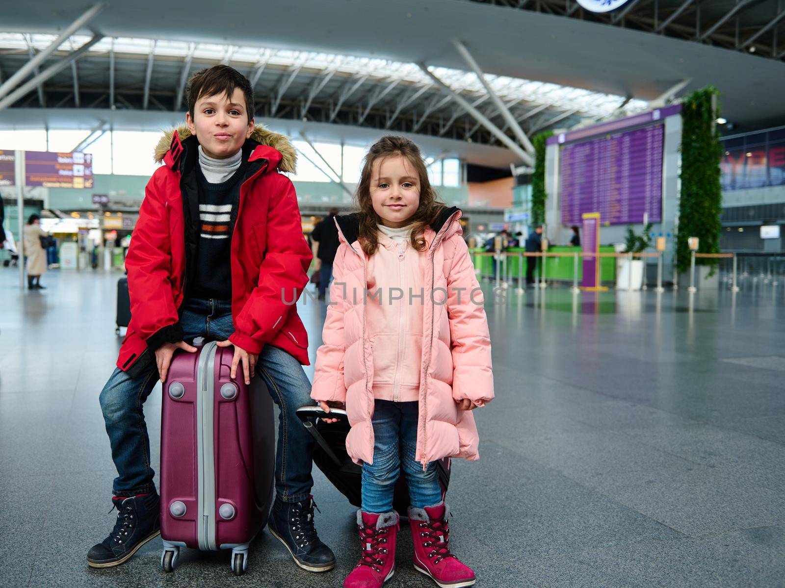 Adorable European children in warm clothes sit on suitcases and look at the camera in the departure area of the international airport. Family holidays,winter tourism and air travel concept. Copy space
