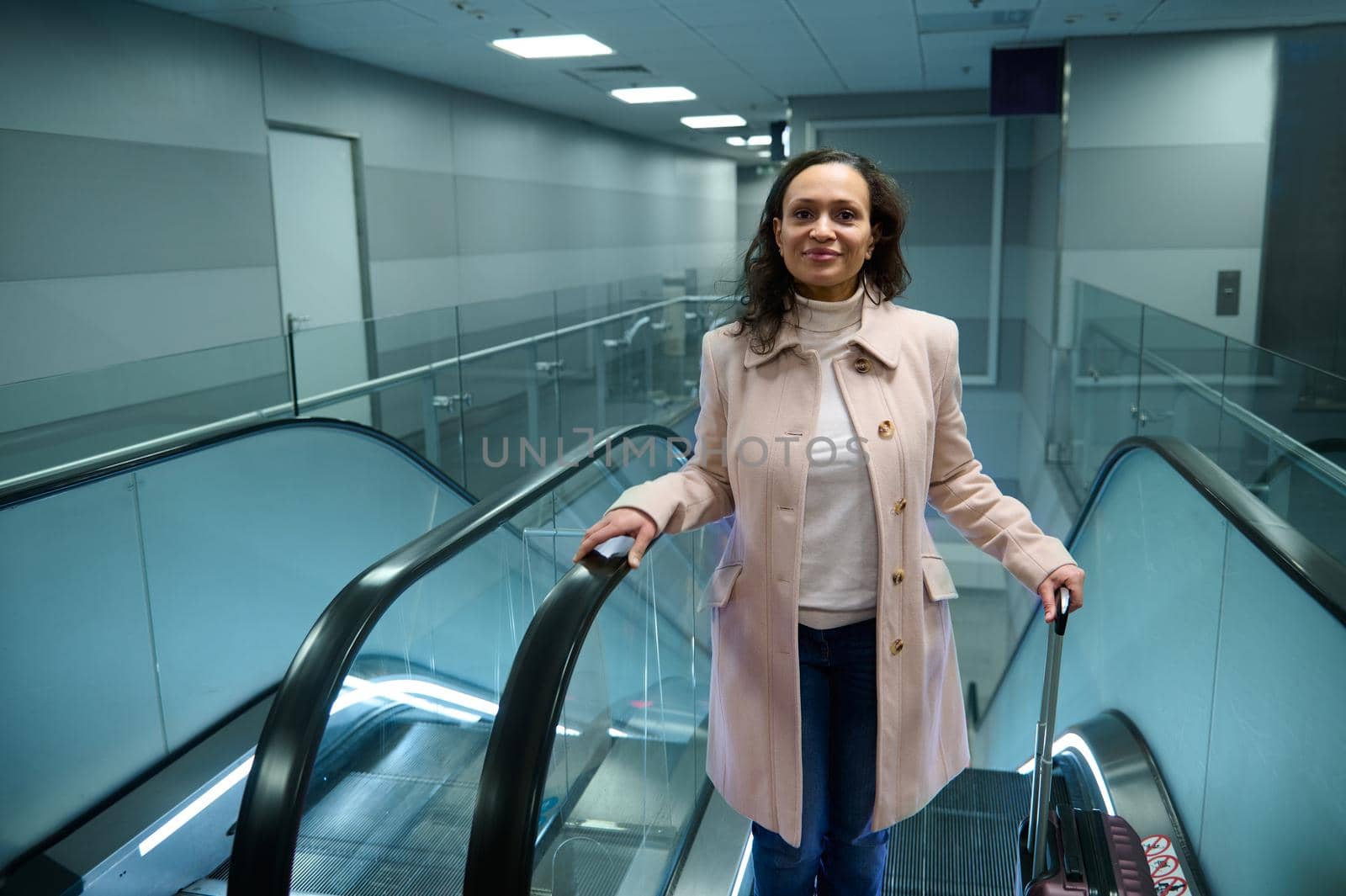 Charming young successful Middle Eastern woman, passenger in casual beige coat holding suitcase and smiling cutely looking at camera while going up escalator in the international airport terminal