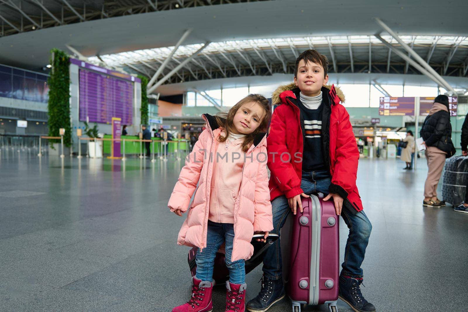 Beautiful elementary age kids, siblings in warm clothes, sitting on suitcases in the departure hall of the international airport, waiting for passenger and customs control and check-in for a flight.