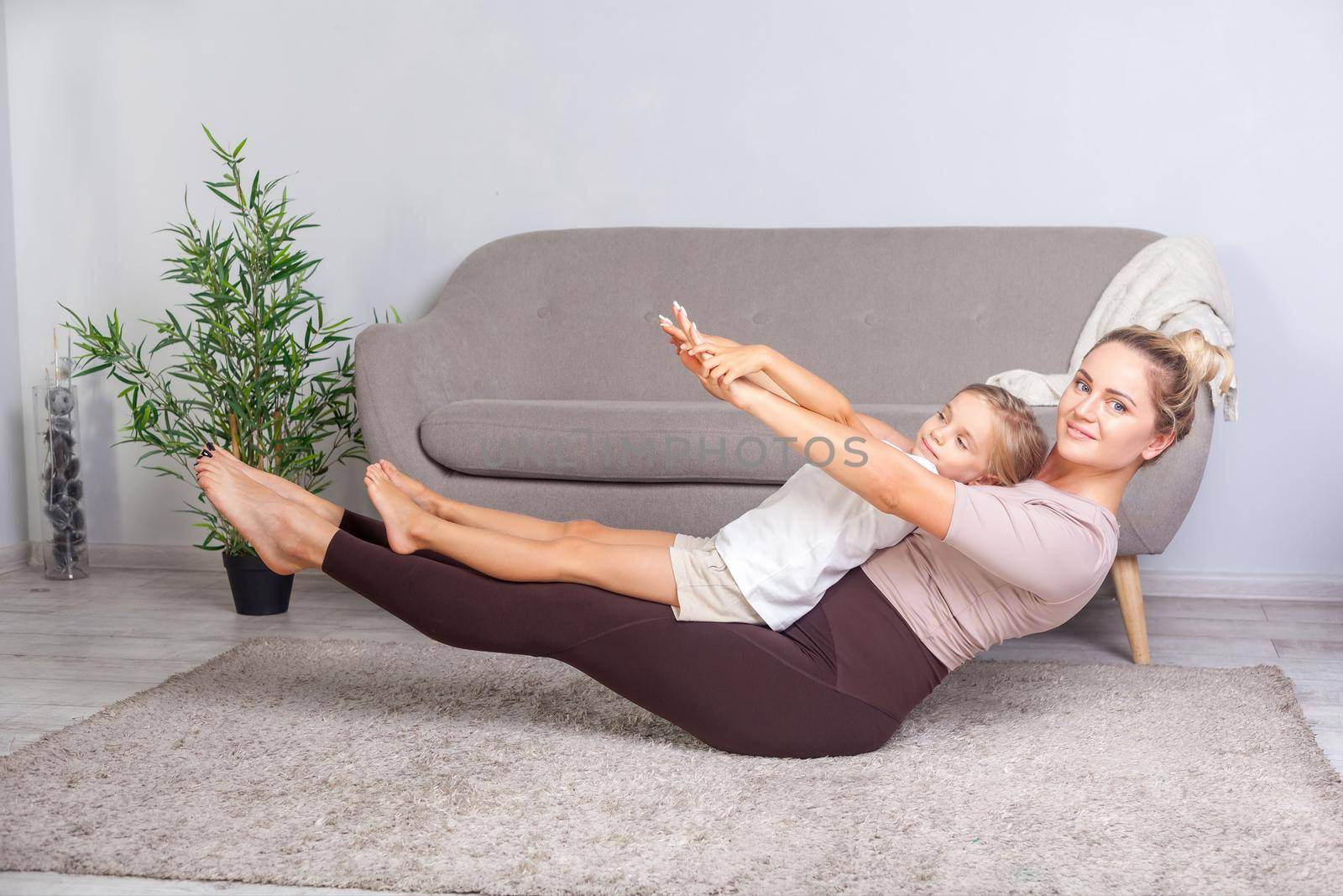 Young beautiful mother and cute daughter practicing yoga together at home, standing in boat pose and looking at camera, happy family doing gymnastics exercise together, fitness with child. healthcare