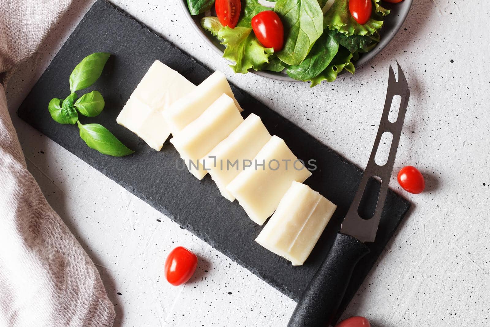 A piece of fresh sliced halloumi on a slate board with cherry tomatoes and spinach leaves. Preparing to cook cheese on the grill. Top view