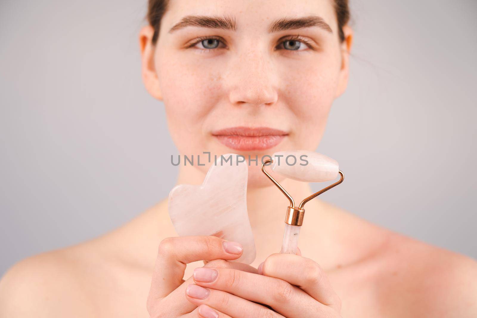 Caucasian woman holding pink roller massager and gouache scraper on white background