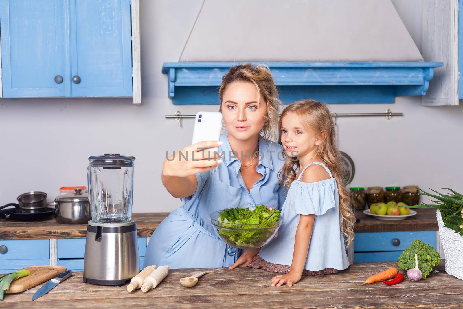 Charming woman and little girl taking selfie in modern kitchen, holding bowl of vegetable salad and posting photo in social network using cell phone, food blog about healthy nutrition, vegetarian diet