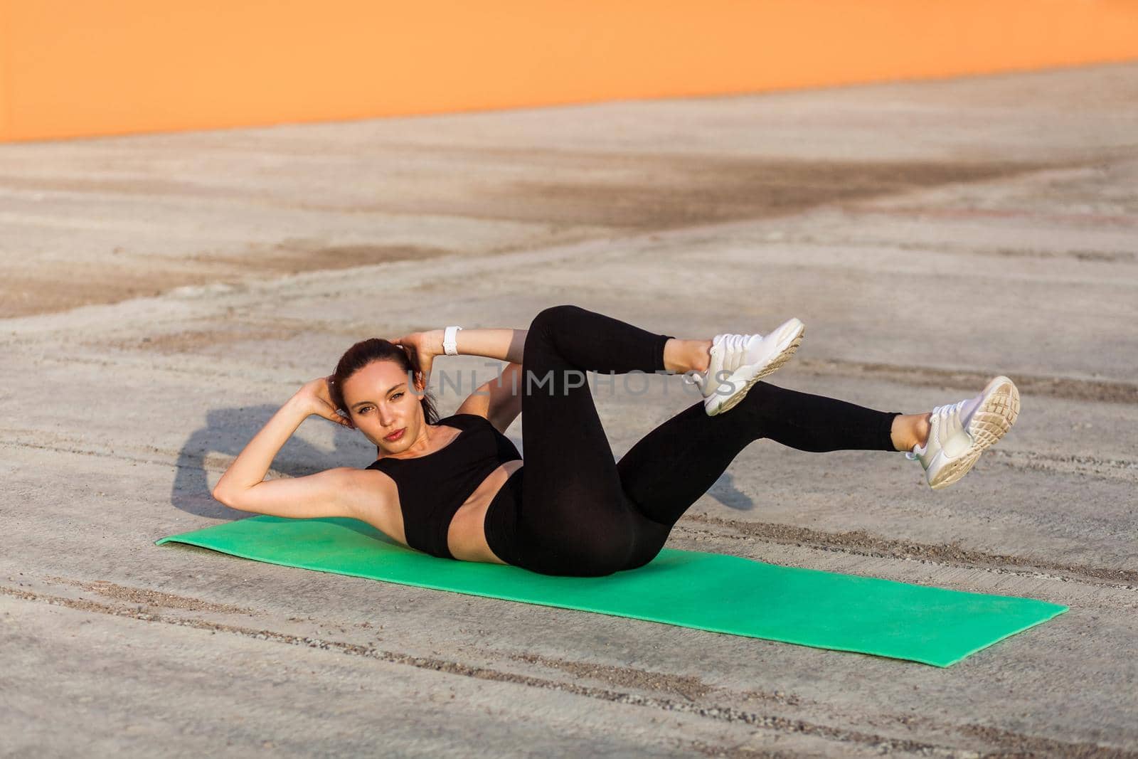 Fit sporty beautiful girl in tight sportswear, black pants and top, lying on mat doing sit ups, crunch exercise, training abdominal muscles. Health care and weight loss concept, sport activity outdoor