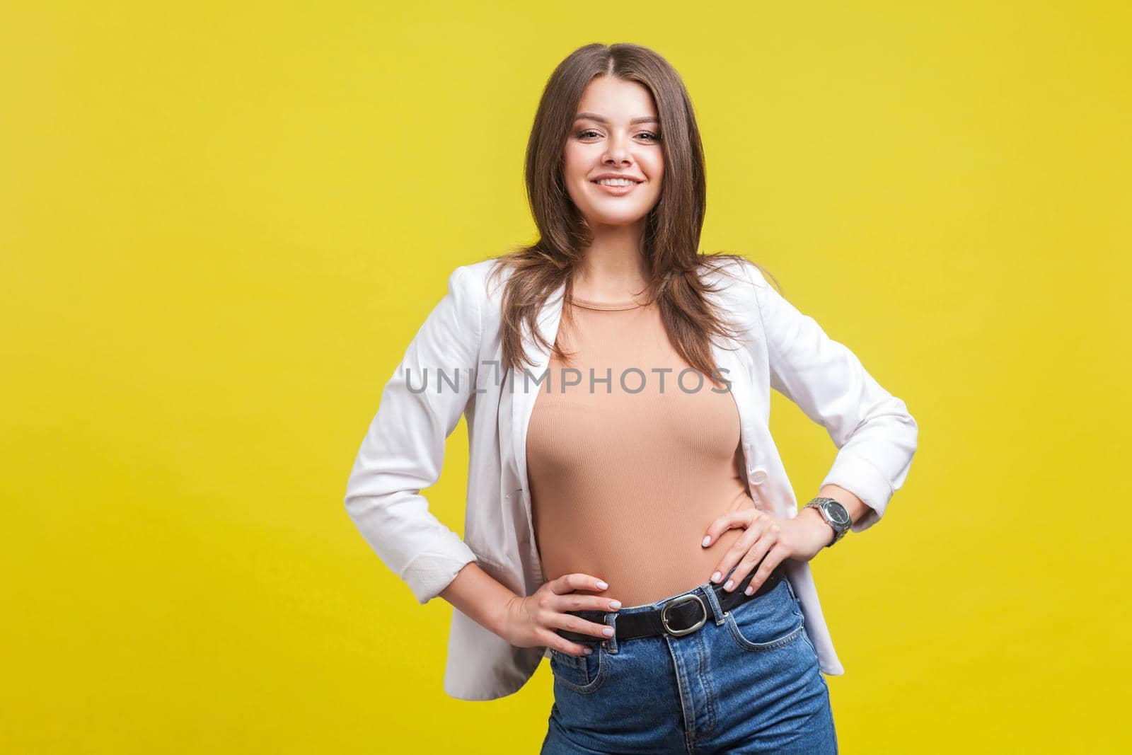 Portrait of stunning self-confident brunette woman in jacket and jeans holding hands on her hips and looking at camera with attractive toothy smile. indoor studio shot isolated on yellow background