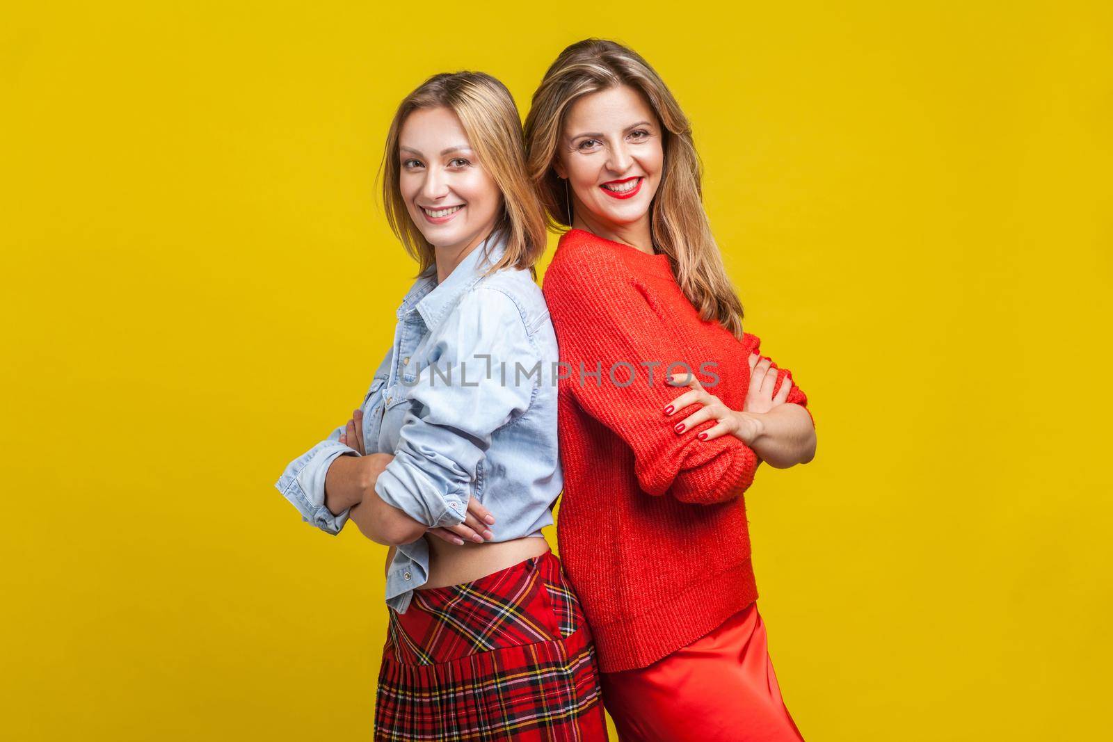 Best friends standing with their backs to each other and crossed hands. Portrait of two charming women in stylish casual clothes smiling at camera. indoor studio shot isolated on yellow background