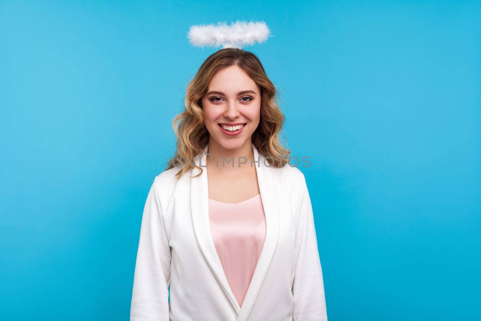 Portrait of gorgeous angelic woman with halo on her wavy hair and in white clothes looking at camera with toothy smile, cute sensual girl with nimbus. indoor studio shot isolated on blue background