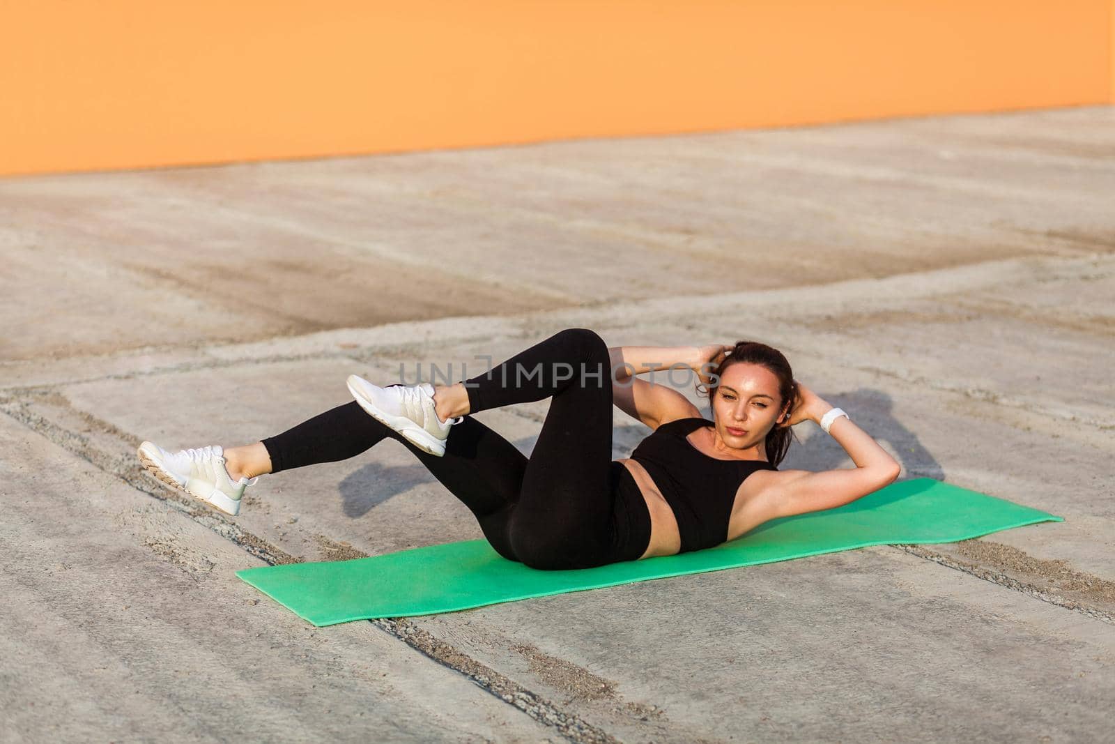 Fit sporty beautiful girl in tight sportswear, black pants and top, lying on mat doing sit ups, crunch exercise, training abdominal muscles. Health care and weight loss concept, sport activity outdoor