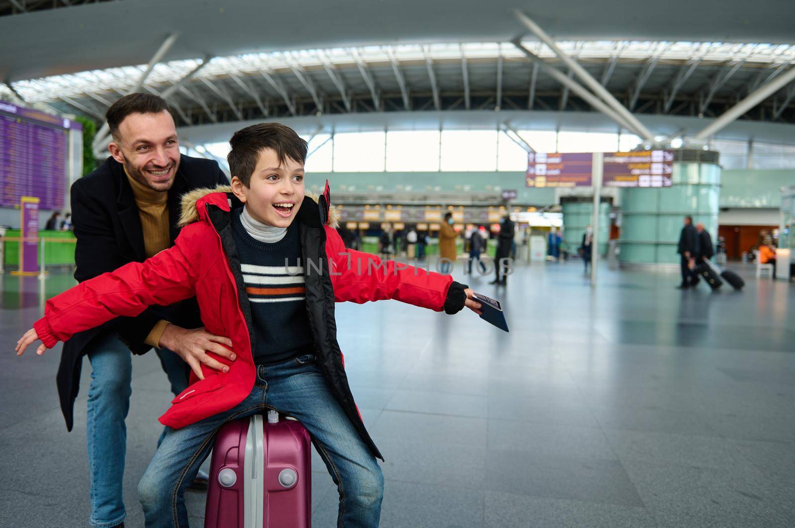 Adorable child boy with passport and boarding pass in his outstretched hands while his cheerful father is riding him on a suitcase in the departure hall of the airport, waiting check-in for a flight