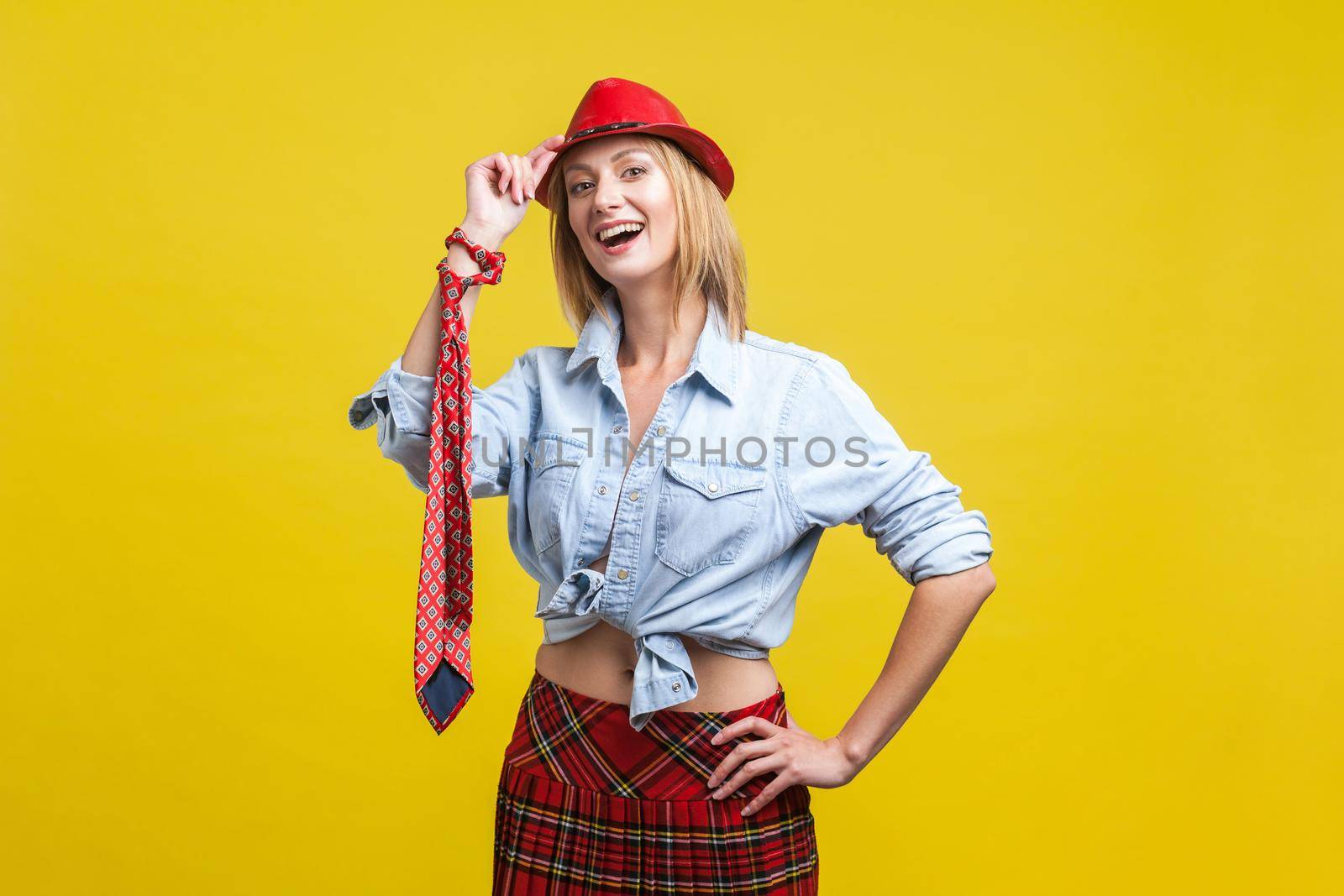 Portrait of charming playful girl looking stylish in denim shirt, school skirt and tie knotted on hand, touching hat on head as if greeting, fashion trend. studio shot isolated on yellow background