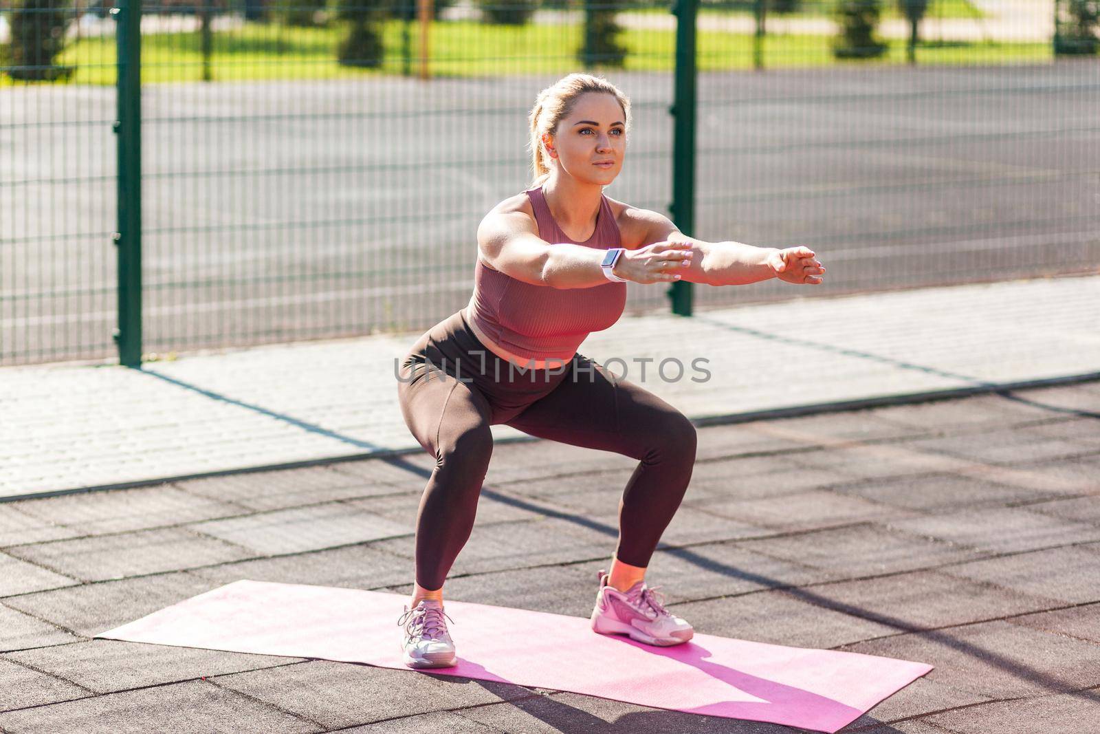 Full length, woman with athletic body in tight pants training on mat outdoor summer day, doing squats with outstretched hands, aerobics or pilates exercise. Health care, sport activity for weight loss