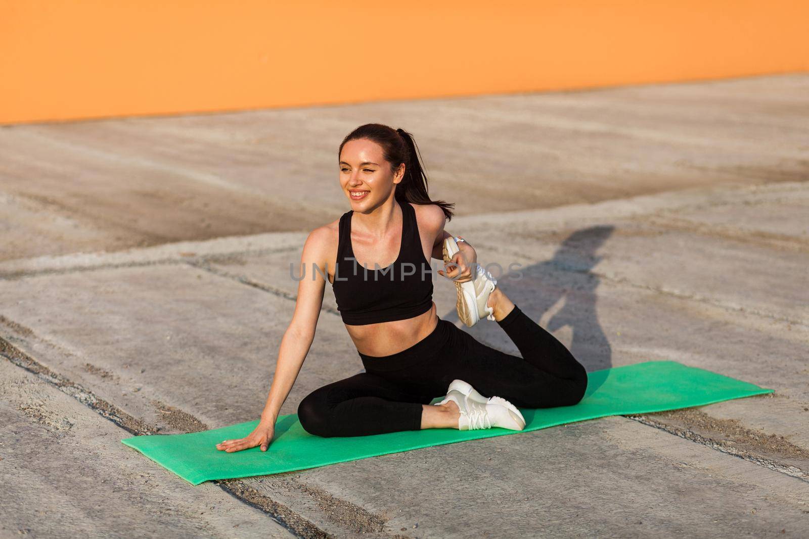 Smiling athletic girl in tight sportswear, black pants and top, practicing yoga, doing one-legged king pigeon pose, stretching leg muscles for better flexibility. Health care, sport activity outdoor