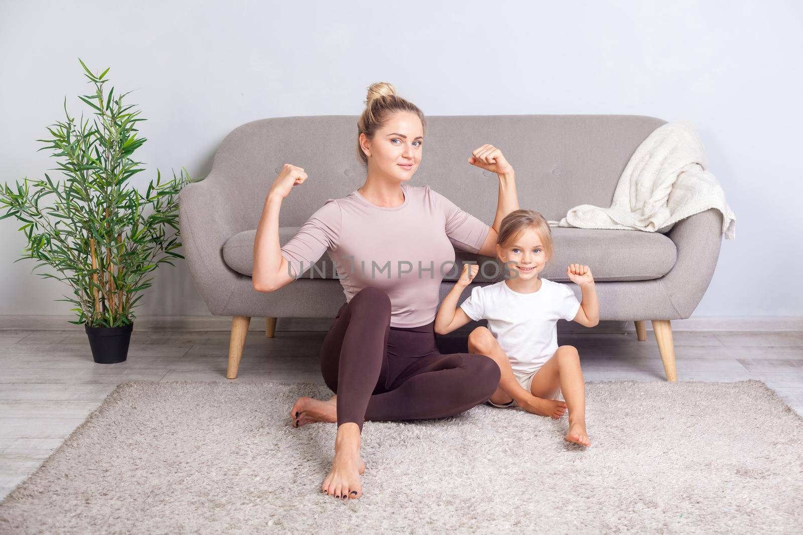 Attractive fit mom and her charming daughter showing biceps, smiling at camera, sitting together on floor after yoga workouts at home, feeling strength and power in trained body. healthcare, sport