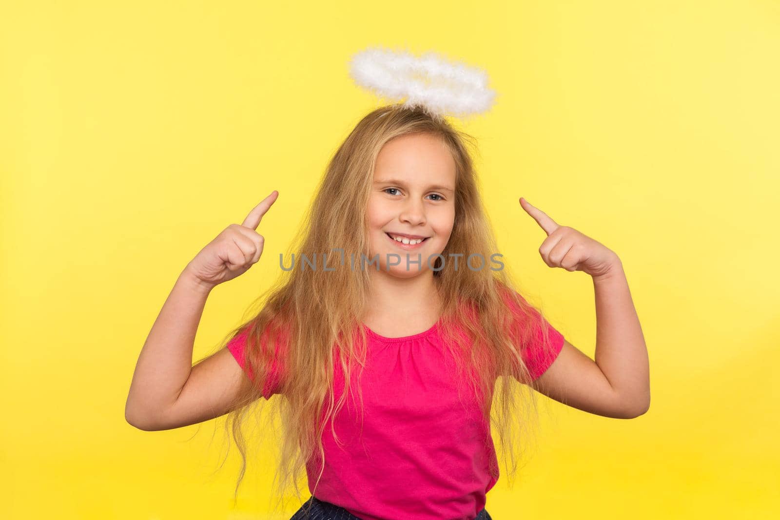 Portrait of adorable happy little girl with long fair hair pointing at angelic halo over head, looking at camera with toothy smile, carefree optimistic childish view. indoor studio shot, isolated