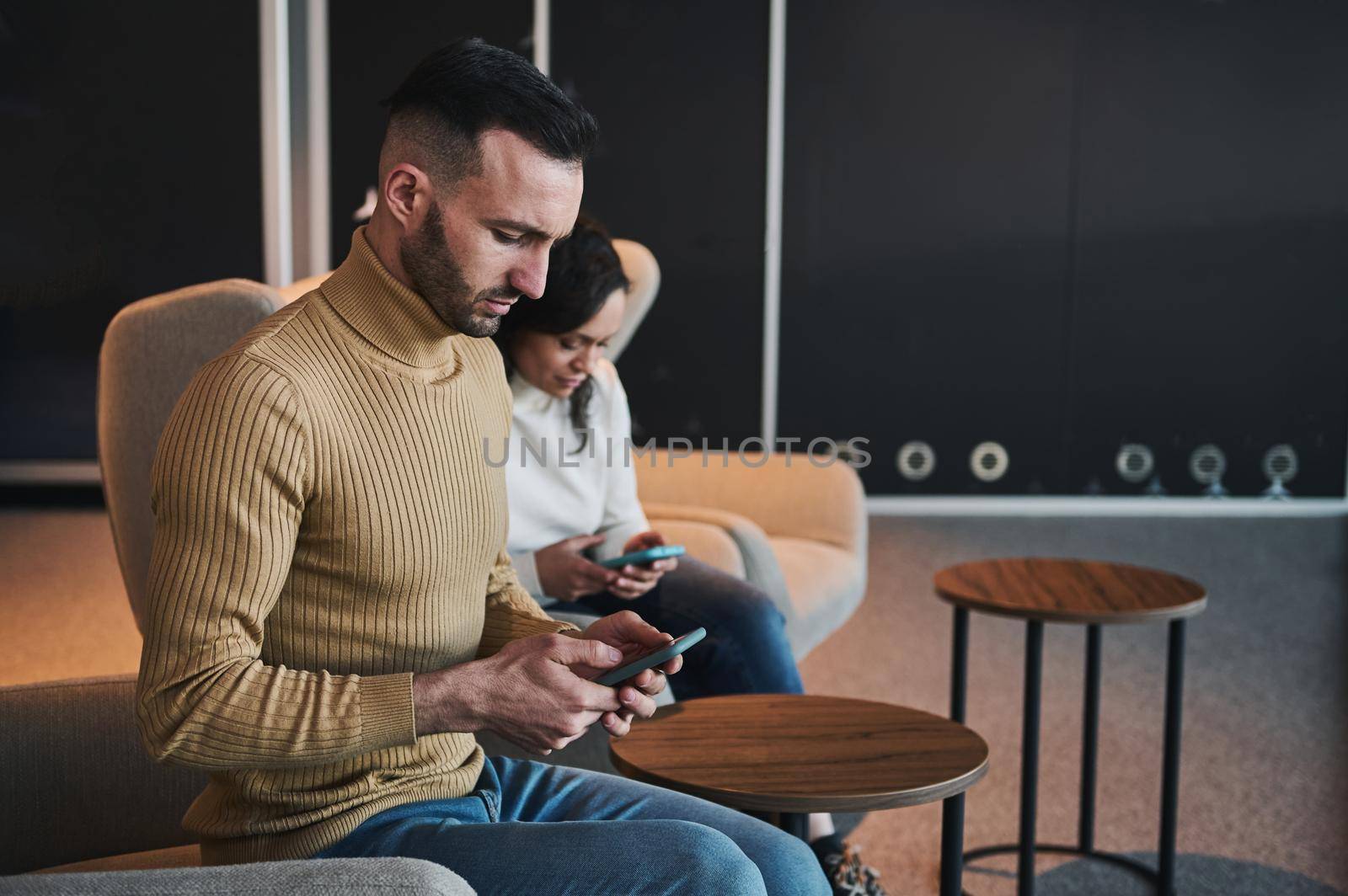 Concentrated multitasking young Caucasian man using mobile phone, booking a hotel, sitting next to his female partner, girfriend, wife in the VIP lounge of the international airport departure terminal