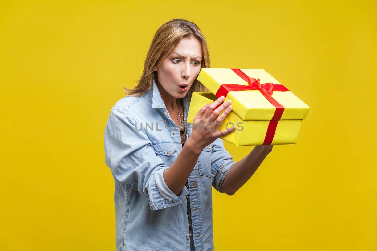 Holiday surprise. Portrait of astonished curious woman in denim shirt looking inside gift box, checking what's inside, unboxing with wow amazed expression. studio shot isolated on yellow background