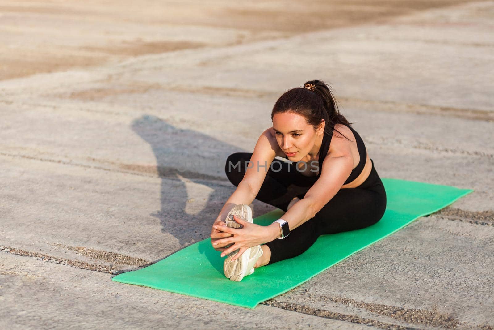 Positive athletic fit woman in tight sportswear, black pants and top, practicing yoga, doing Head-to-Knee Pose, touching toes, stretching leg and back muscles. Health care, sport activity outdoor