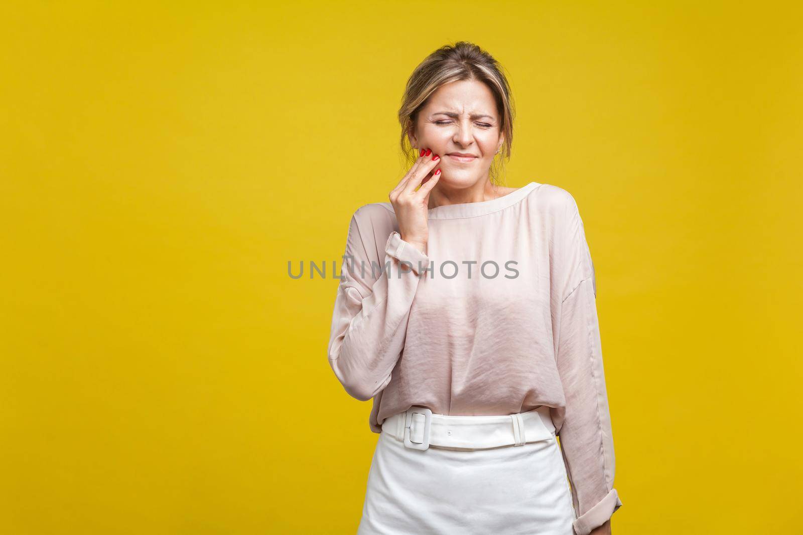 Portrait of upset sick young woman with fair hair in casual blouse standing with closed eyes, touching cheek suffering dental teeth ache, mouth injury. indoor studio shot isolated on yellow background